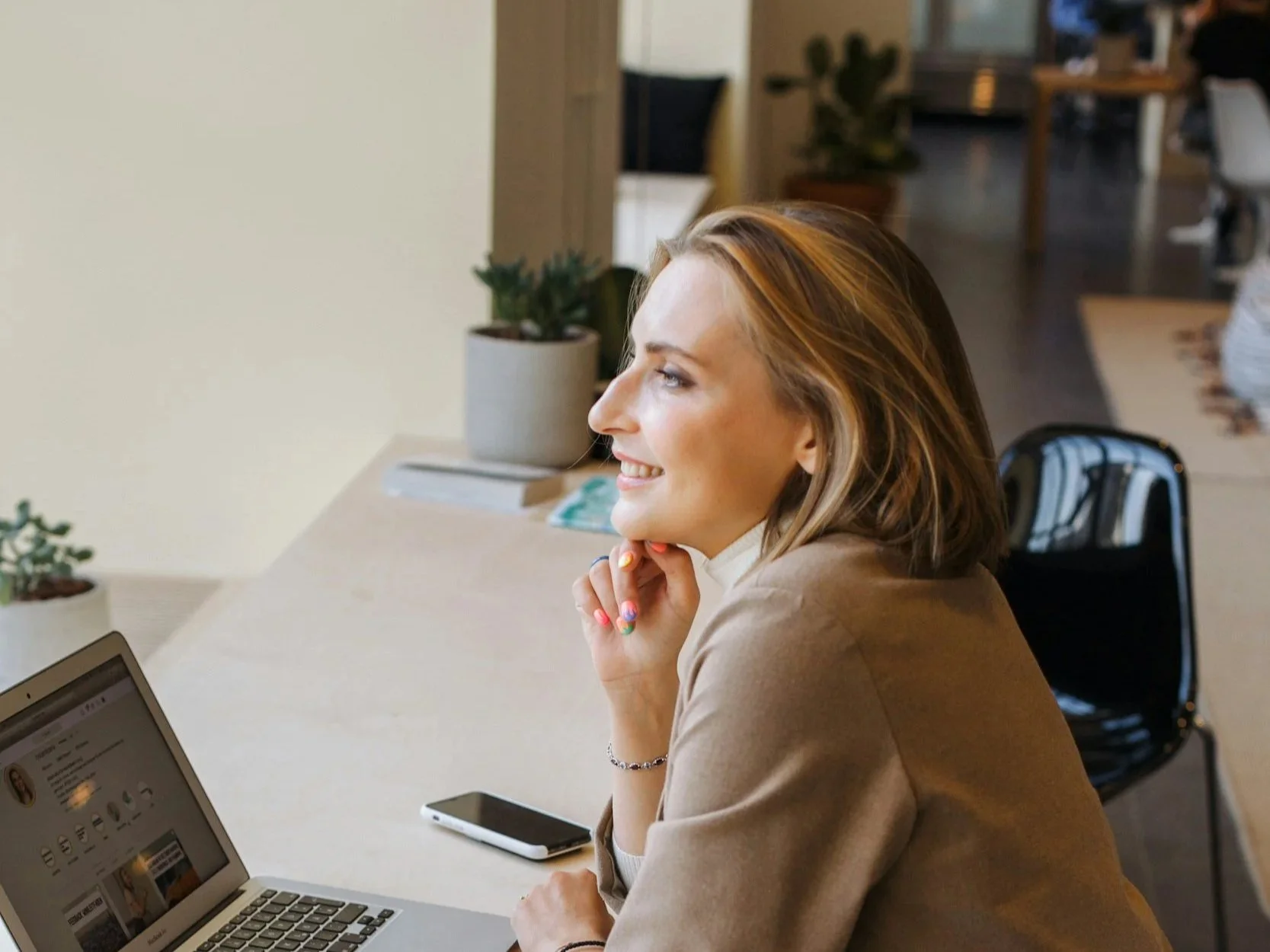 A woman with shoulder-length hair smiling while sitting at a desk with a laptop and a smartphone. She is in an office setting with some potted plants and books in the background.