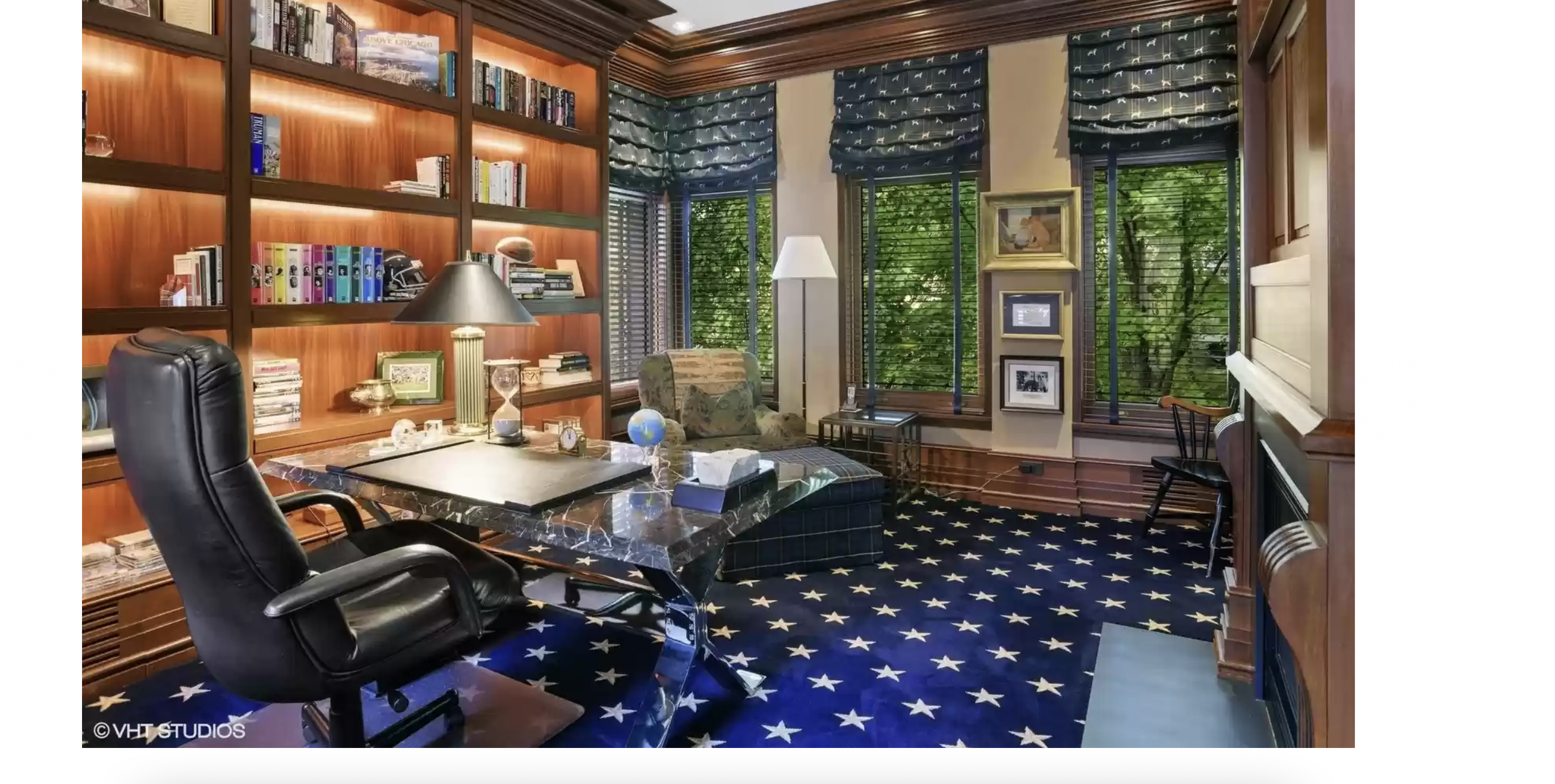 A home office with wooden walls, a dark desk with a black leather chair, a patterned armchair, tall bookshelves filled with books, and windows with blue patterned curtains showing green trees outside.