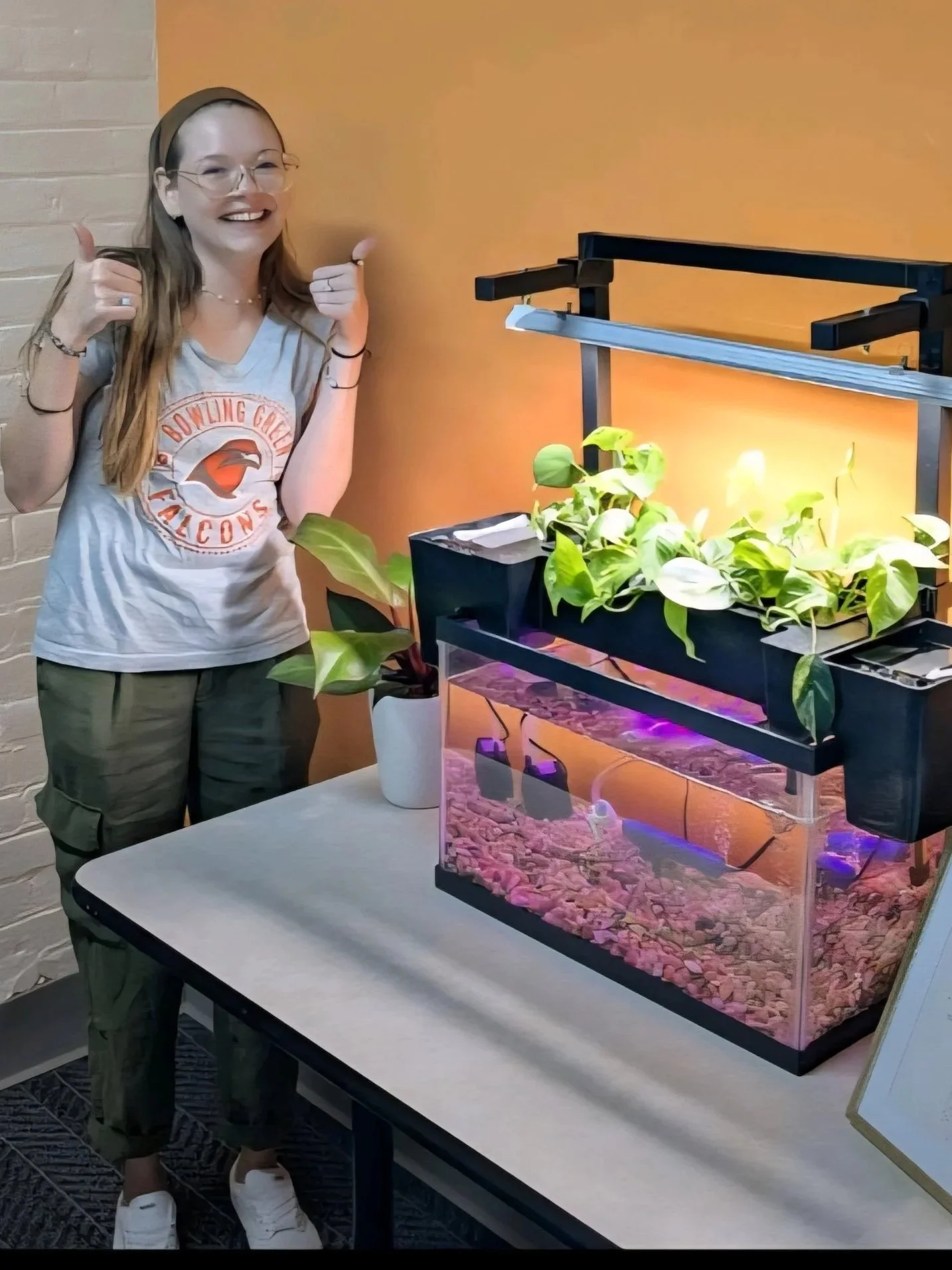 Our Founder: A young woman with long hair, glasses, and a gray t-shirt, standing next to a terrarium with plants inside and giving two thumbs up.