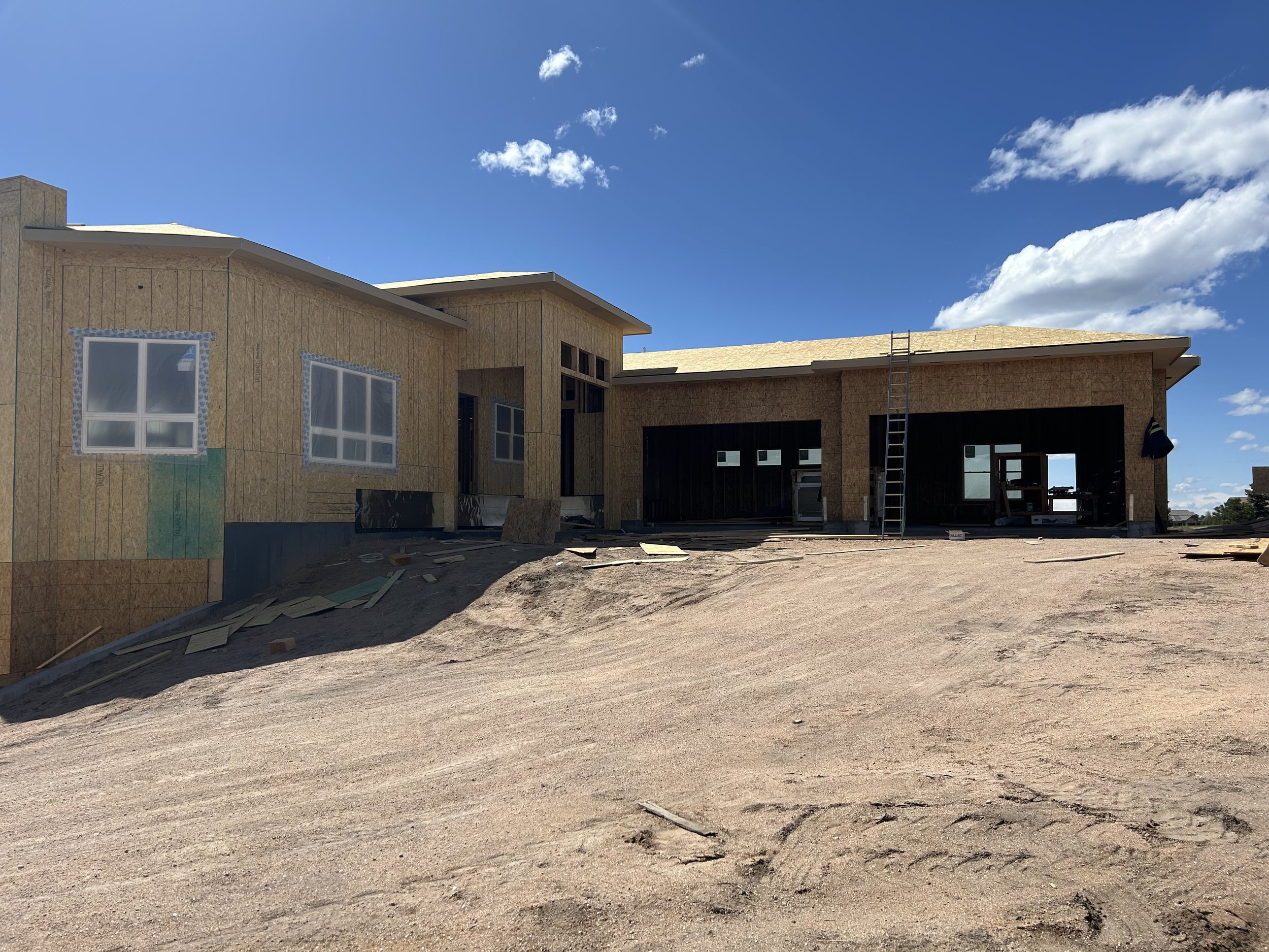A house under construction with unfinished wood siding and a partly open garage, with construction materials scattered on dirt ground under a blue sky with clouds.