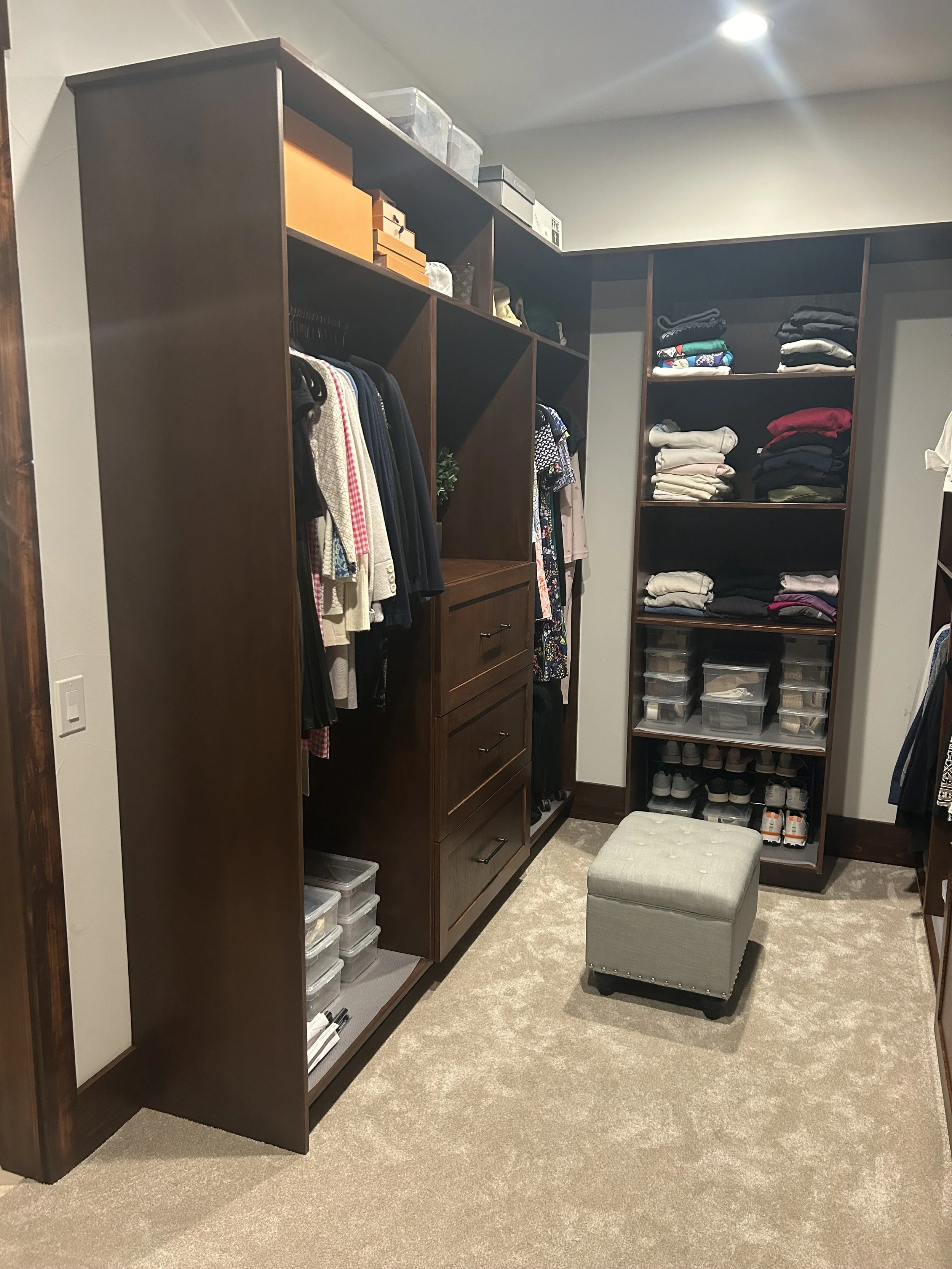 A walk-in closet with dark wood shelving units filled with folded clothes, shoe boxes, and hanging garments, a beige ottoman in the center, and a light-colored carpeted floor.