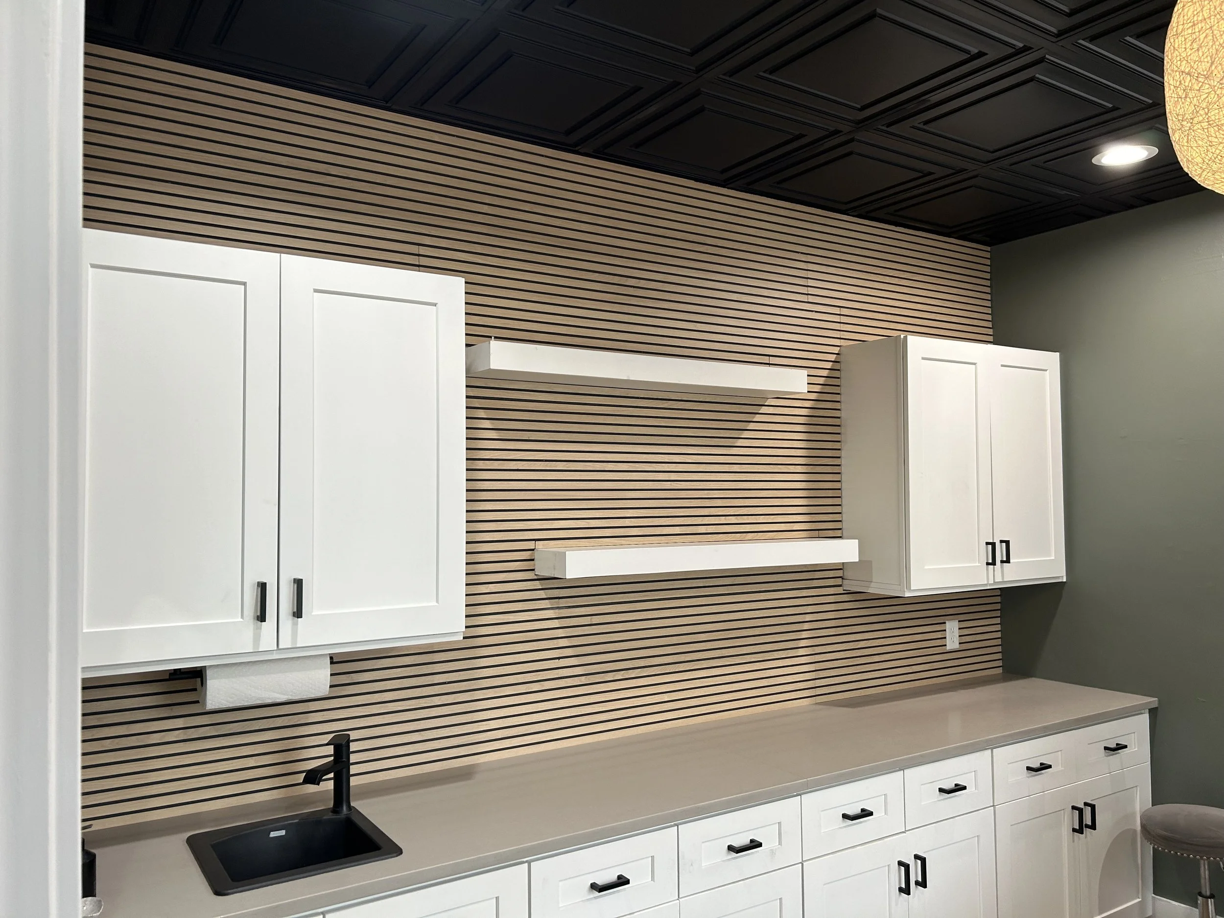Modern kitchenette with white upper cabinets, black faucet, beige countertop, and striped wooden wall accent.