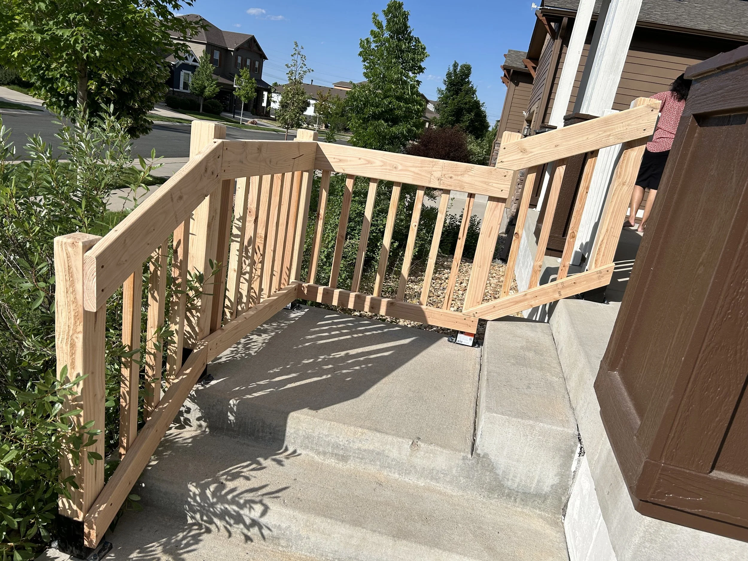 New wooden staircase railing being installed on a porch in a suburban neighborhood with houses, trees, and clear blue sky.