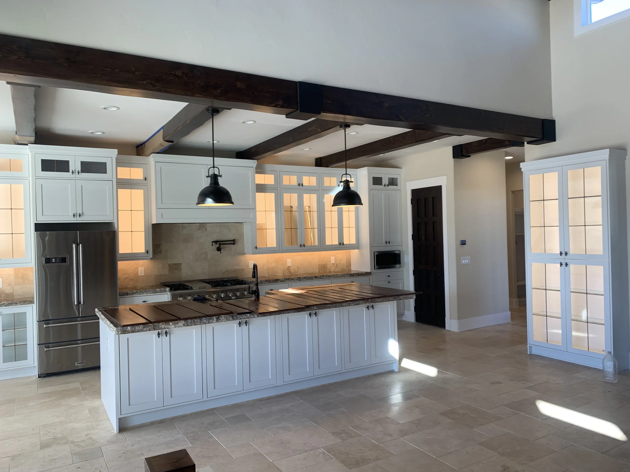 Modern kitchen with white cabinets, a stainless steel refrigerator, a central island with a dark wooden top, black pendant lights, and a dark wooden door on the wall. The kitchen has beige tile flooring and lots of natural light.