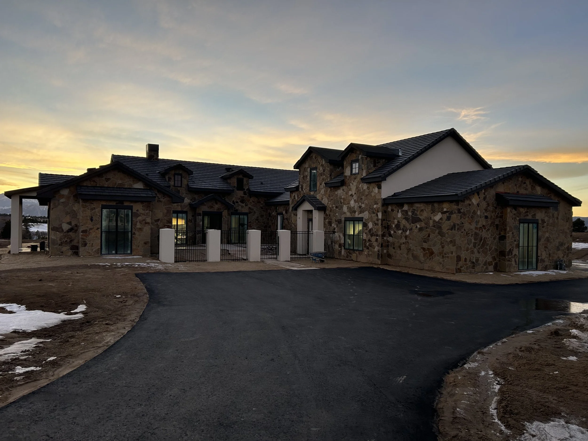 A newly constructed house with stone exterior walls and a black metal roof at sunset, with a paved driveway leading up to it and patches of snow on the ground.