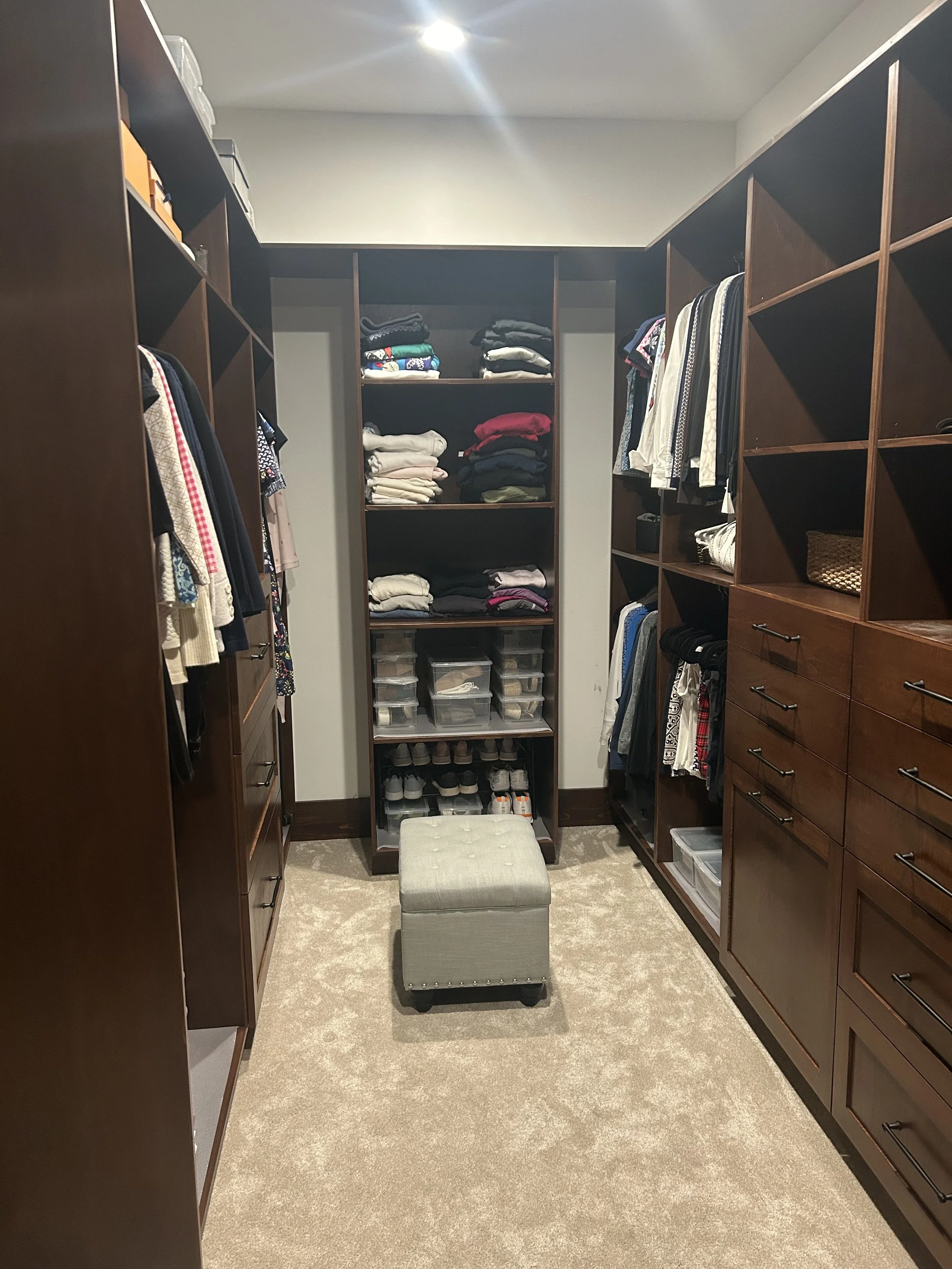 A walk-in closet with dark wood shelving and drawers, filled with folded clothes, shoes, and storage bins. A small, light-colored ottoman sits in the center on a beige carpet.