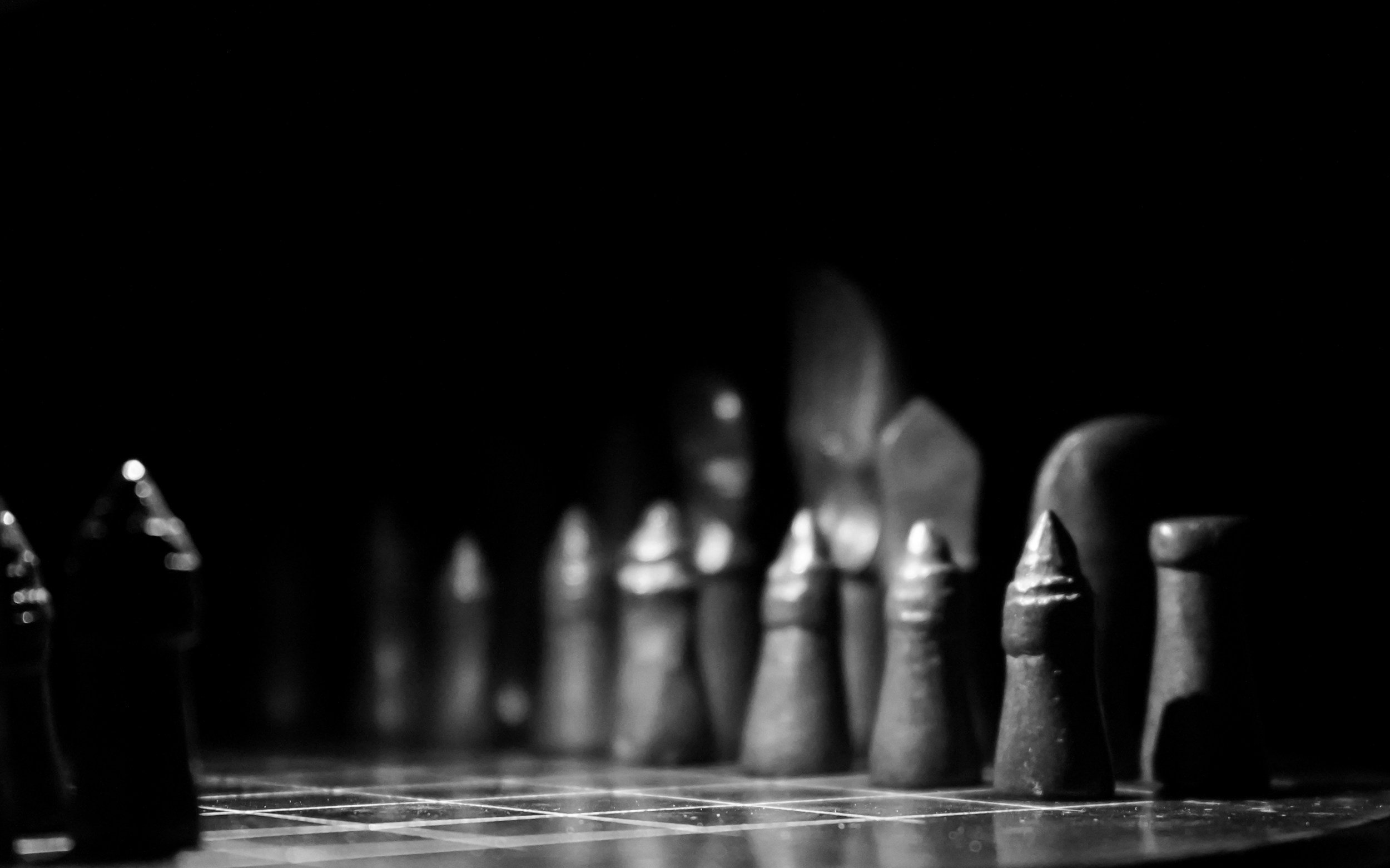 Close-up of a chessboard with black chess pieces in the foreground, shot in black and white with a dark background.
