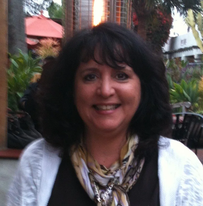 A satisfied customer with dark hair smiling outdoors, wearing a black top, white cardigan, and a patterned scarf, with greenery and a building in the background.
