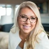 A smiling woman with glasses and long blonde hair, wearing a white shirt, in an indoor setting.