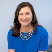 A happy client with shoulder-length brown hair wearing a blue top and layered necklaces, smiling against a light blue background.