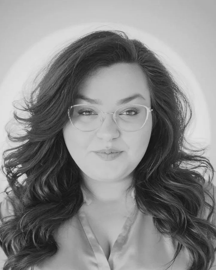 Black and white close-up portrait of a woman with long, wavy hair, glasses, and a confident expression, wearing a satin or silk top.