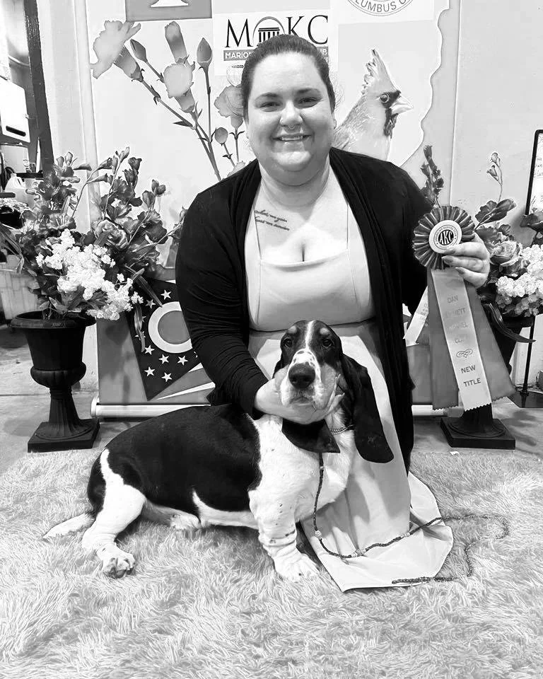 Woman kneeling with a black and white Basset Hound on a red shaggy rug, holding a blue and white ribbon. Background with red, white, and blue decorations, flowers, and a logo, indicating an award ceremony or competition.