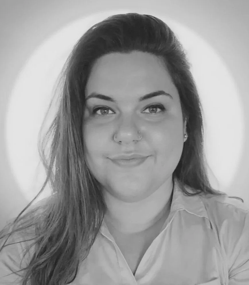 Black and white portrait of a woman with long hair, smiling, with a nose piercing, wearing a collared shirt, with a light source creating a halo effect behind her head.