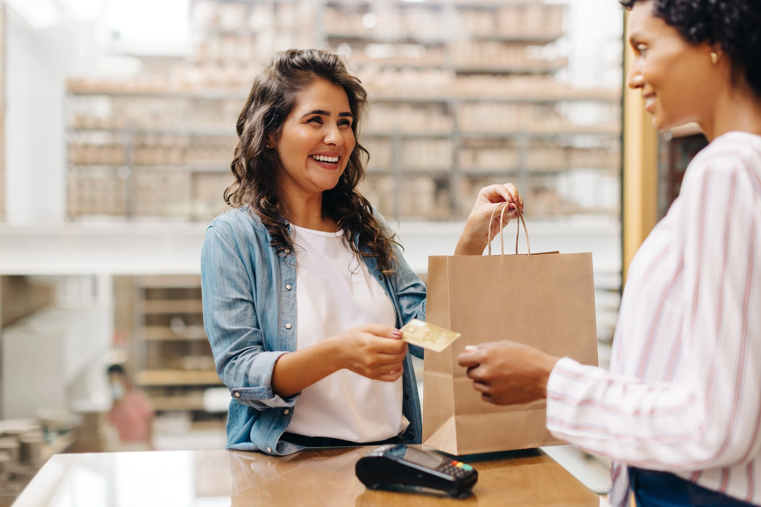 A woman with curly hair smiling as she hands over money in exchange for a paper shopping bag to a cashier in a retail store.