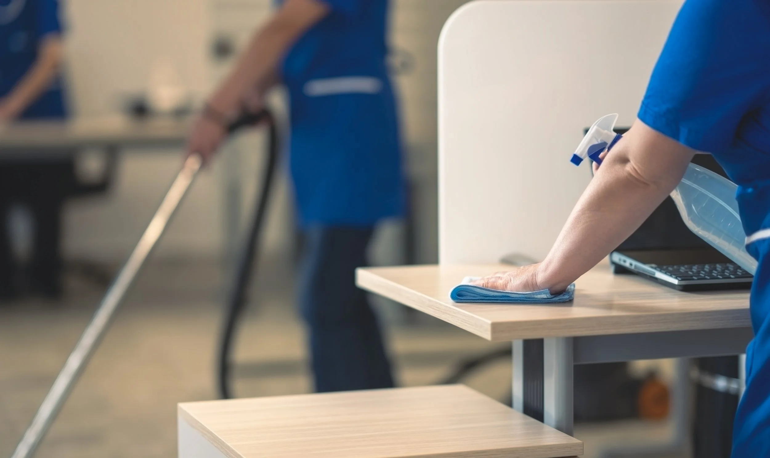 A healthcare worker disinfecting a table with a cloth, with another person in the background using a walking cane in a medical or healthcare setting.