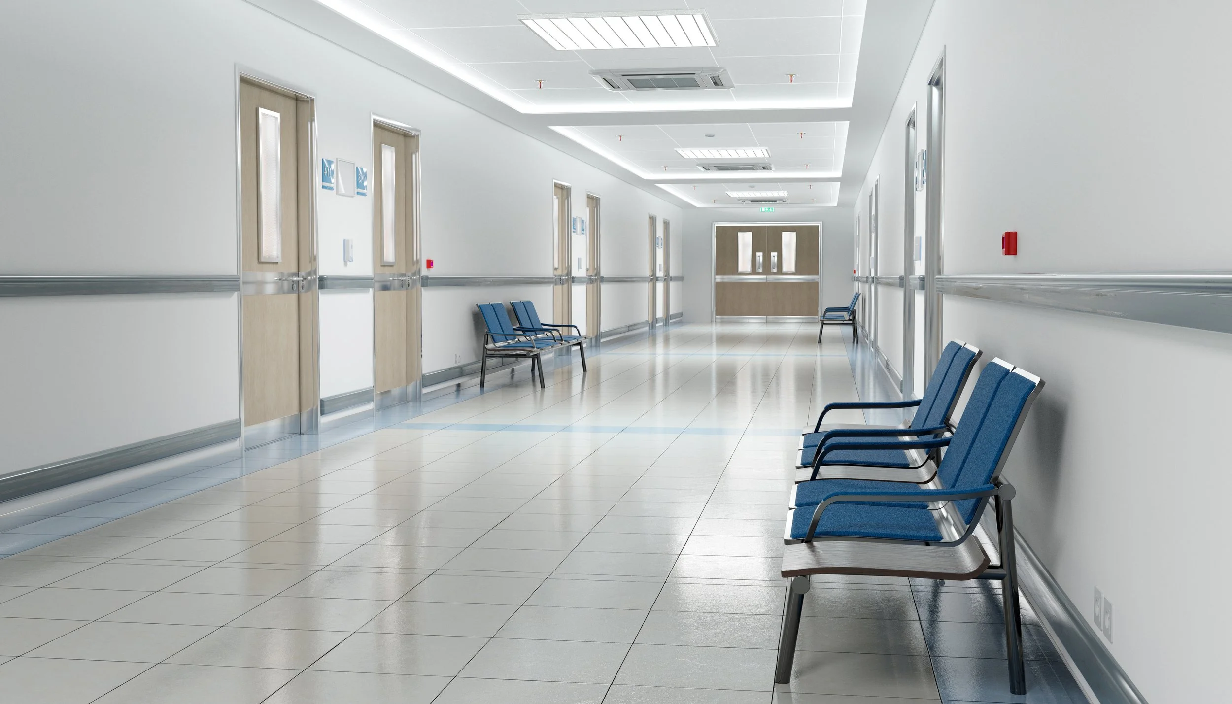 Empty hospital corridor with blue chairs lined along the wall and doorways on both sides.
