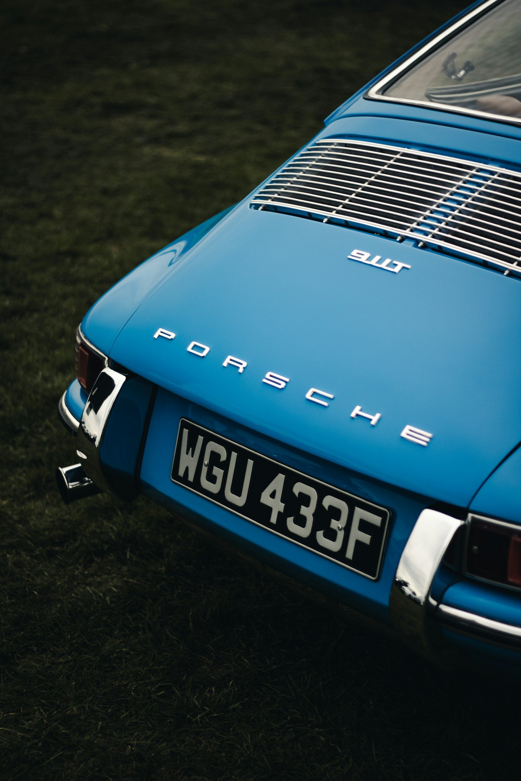 Close-up of a vintage blue Porsche 911 sports car, showing the rear with the Porsche emblem, model 911, and the license plate WGU 433F, on grass.