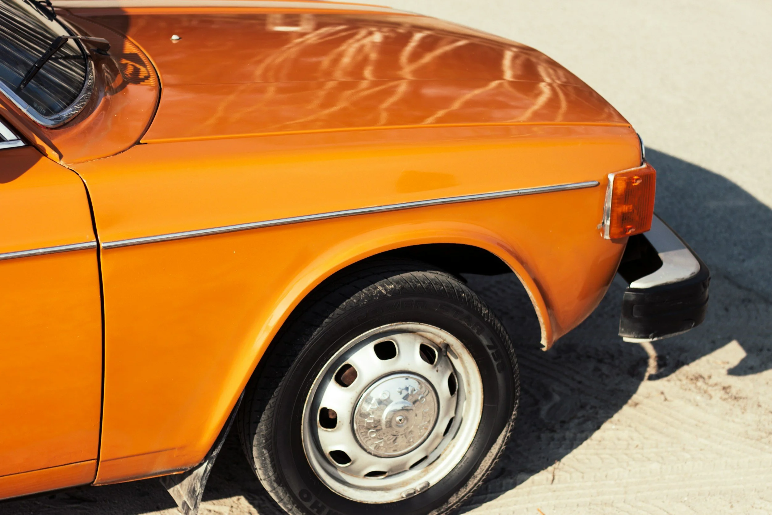 Close-up of the front left side of an orange vintage car, showing the wheel, bumper, and part of the hood, with sunlight reflections on the surface.