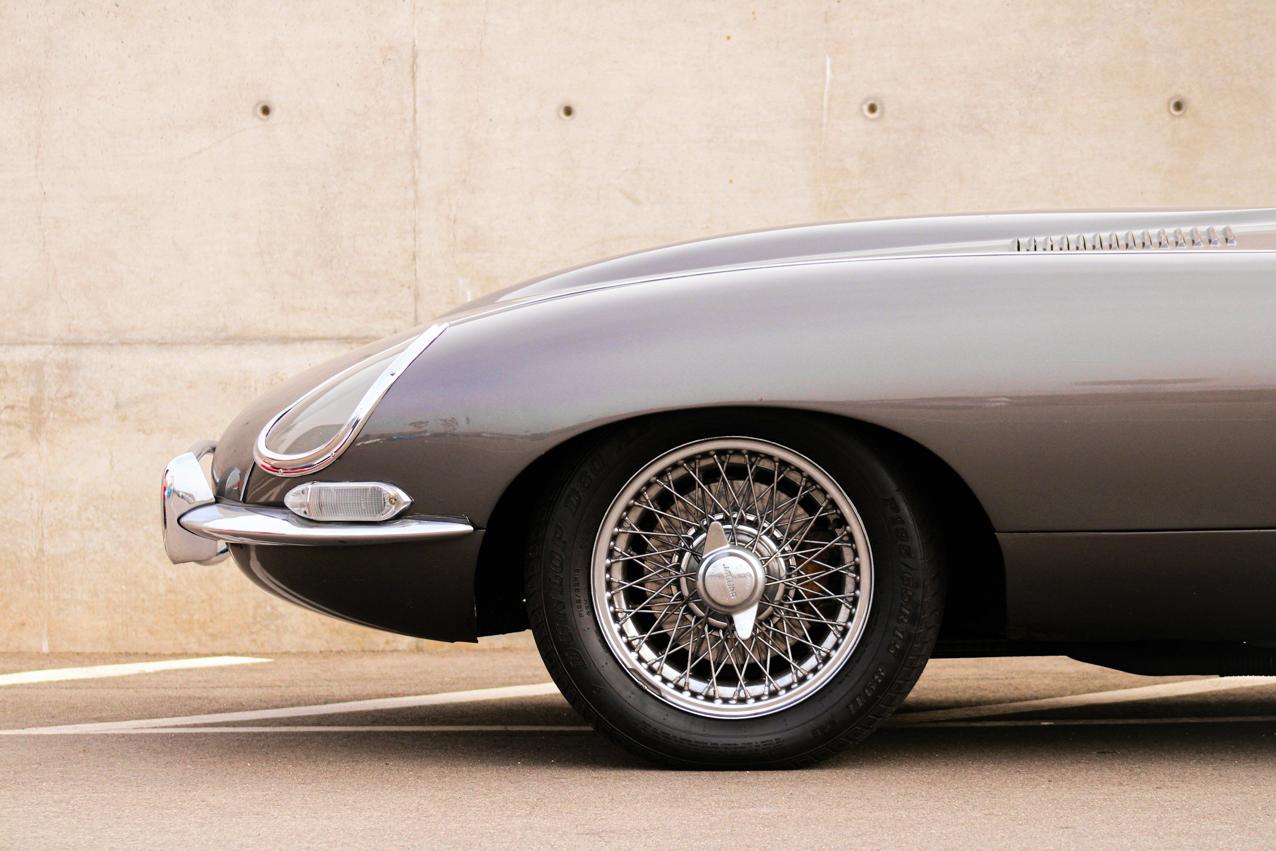 Close-up of the front side of a Jaguar E-type sports car with wire-spoke wheels against a beige concrete wall.