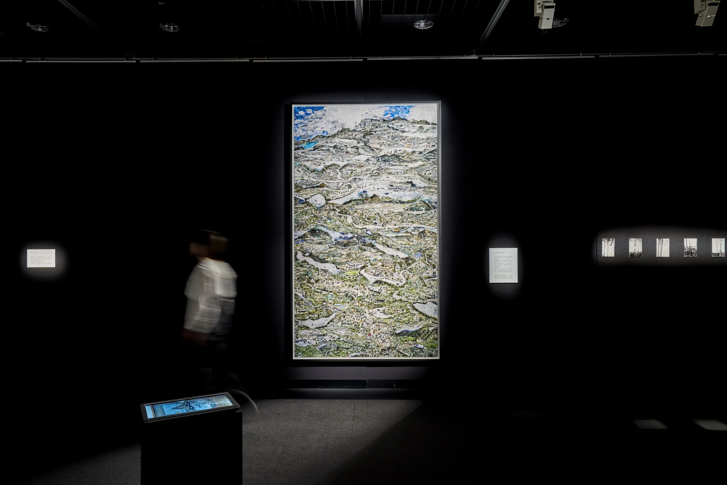 暗い展示室に、大きな縦長の絵画といくつかの解説板、スクリーンが配置されている。