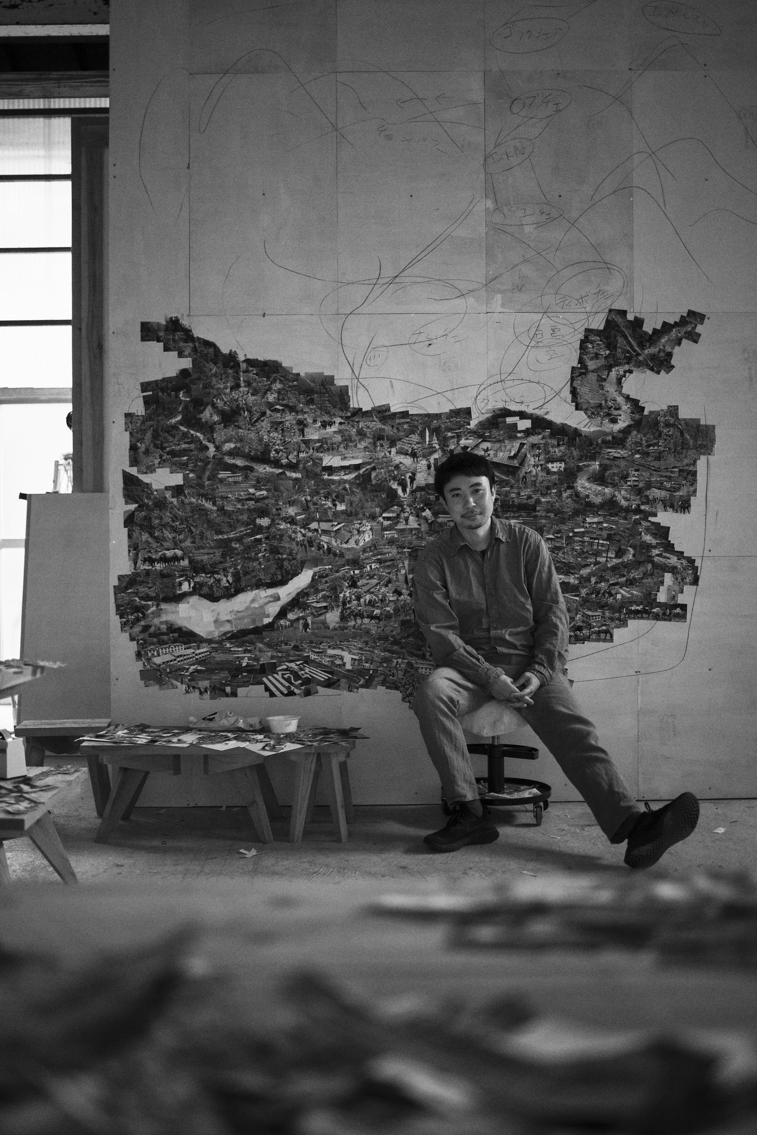 白黒の写真。男性が椅子に座り、背後の壁に大きなコラージュと手書きのスケッチが描かれた壁の前にいます。