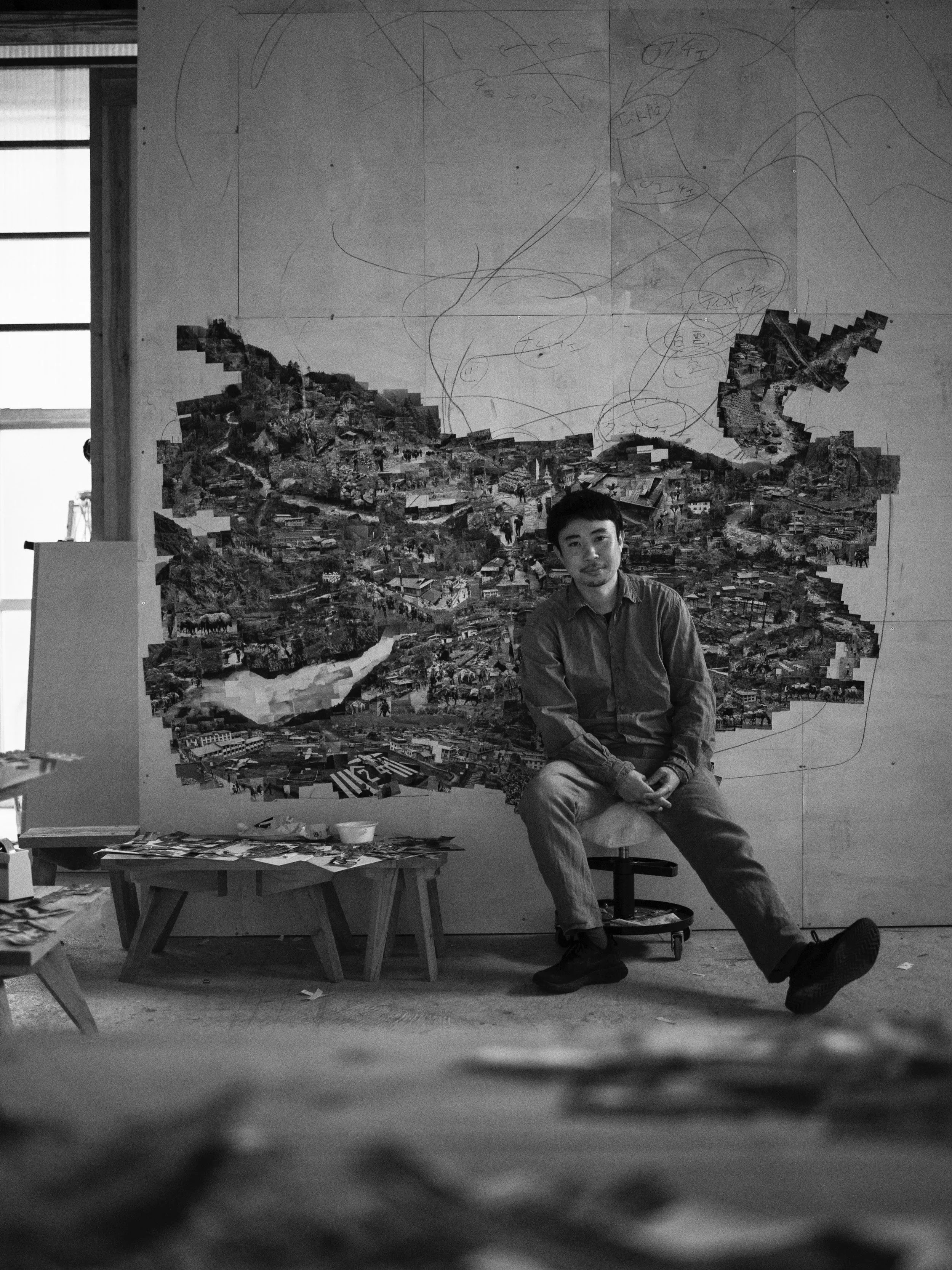 白黒の写真で、男性がうつっている。背景には都市の地図のコラージュとスケッチが描かれた壁がある。男性は椅子に座り、カメラを見ている。