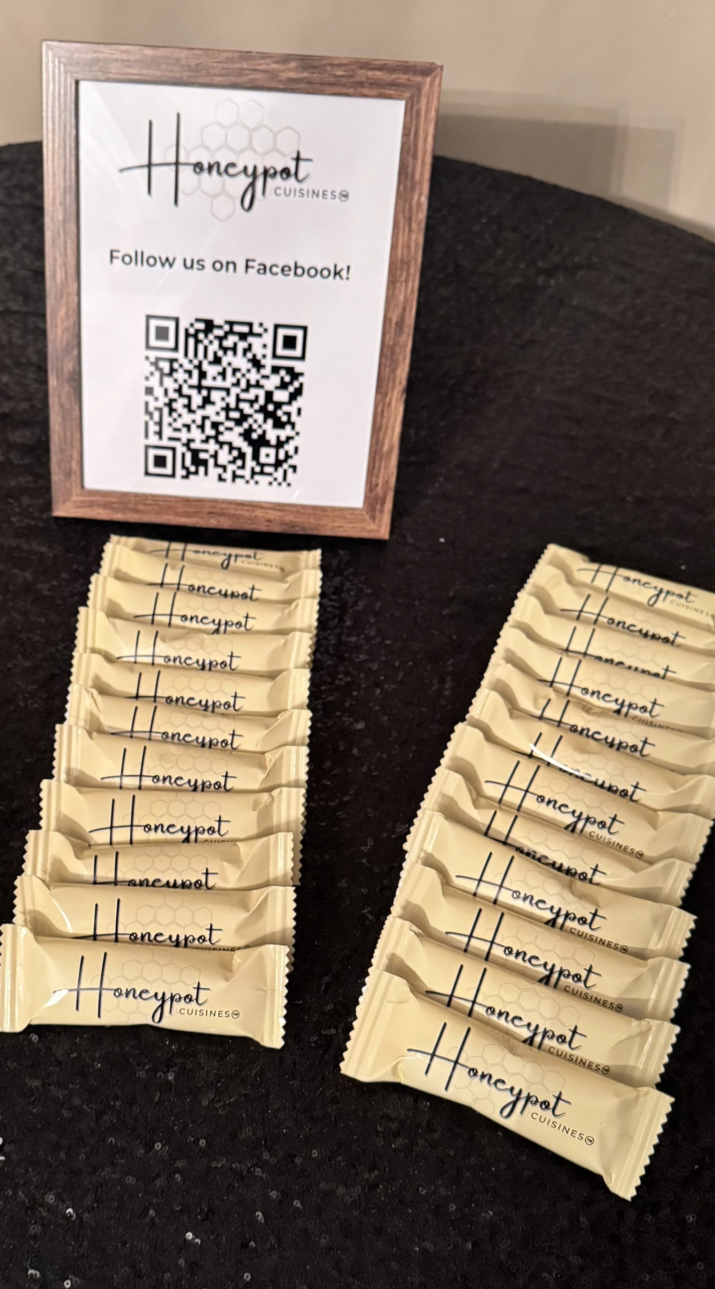 Table display with Honey Pot Cuisine branded candy bars arranged in two groups and a frame with a QR code for Facebook linking on a black tablecloth.