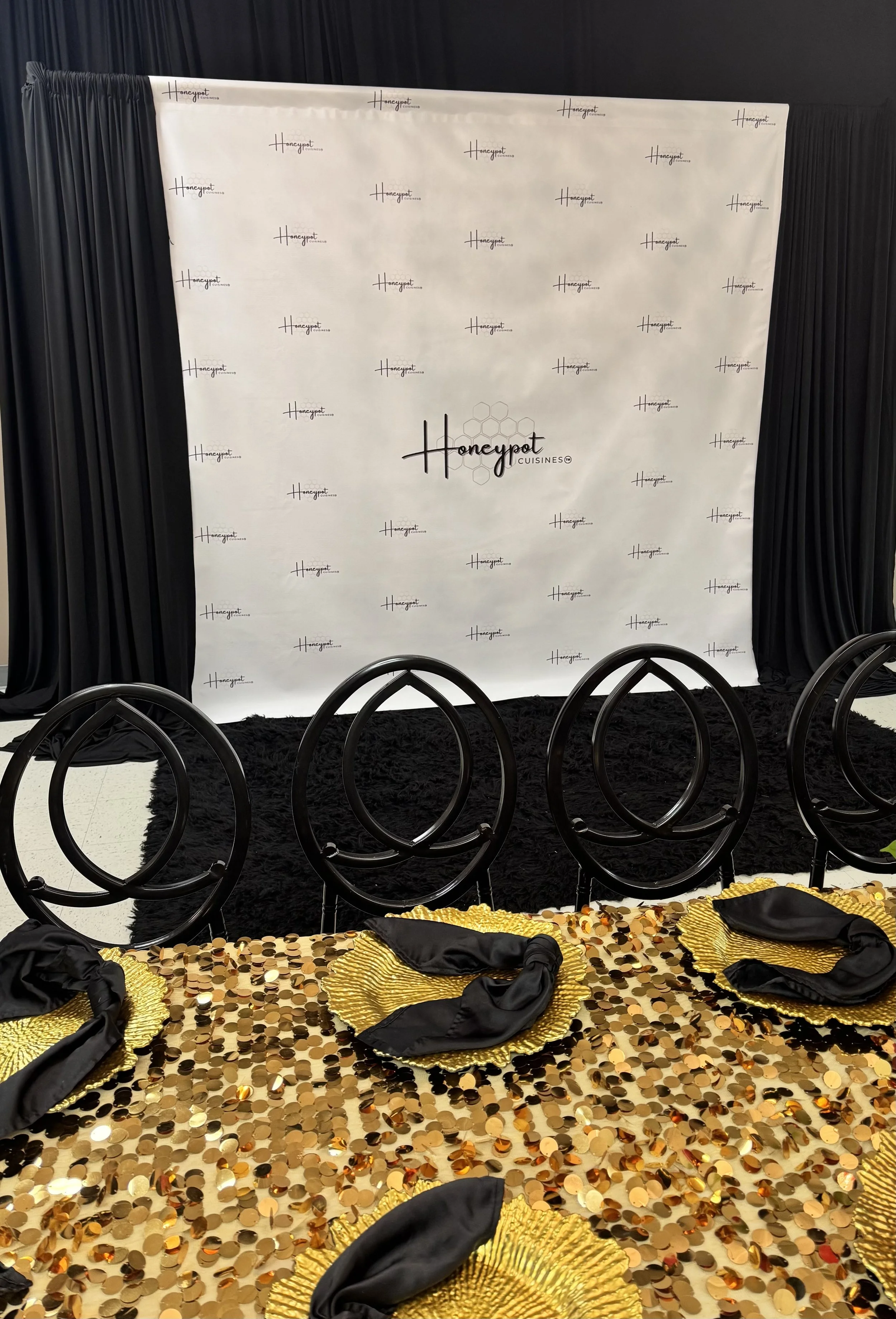 Event decor with a gold and black color theme, featuring a backdrop with 'Honeypot Cuisines' logos, a black curtain, and a table with a gold sequin tablecloth, black napkins, and black chairs.