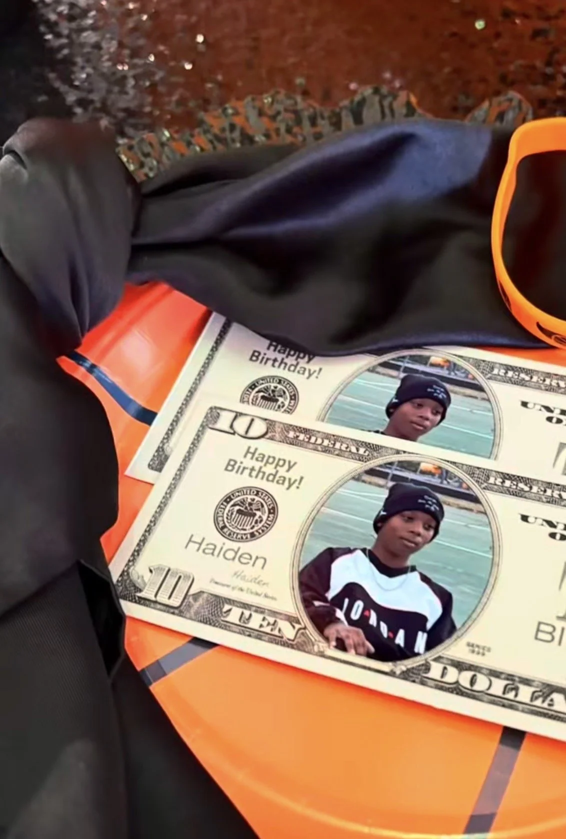 Two ten-dollar bills on an orange surface, each featuring a photo of a young boy wearing a black beanie, with the caption "Happy Birthday!" and the name "Haiden." The surrounding items include a pair of orange sunglasses and black clothing.