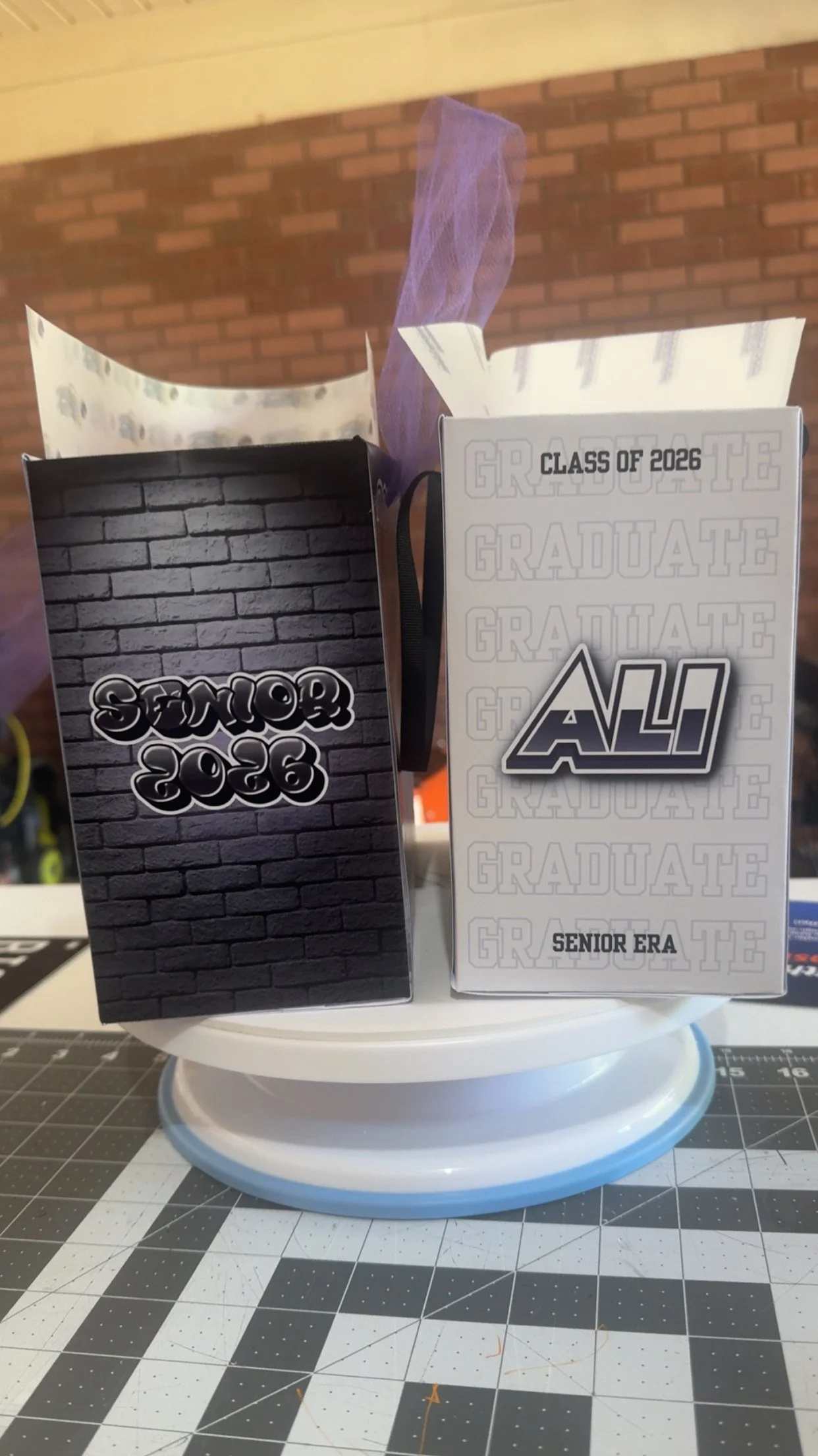 Two graduation gift boxes on a white display stand, with a brick wall background and a cutting mat surface below.