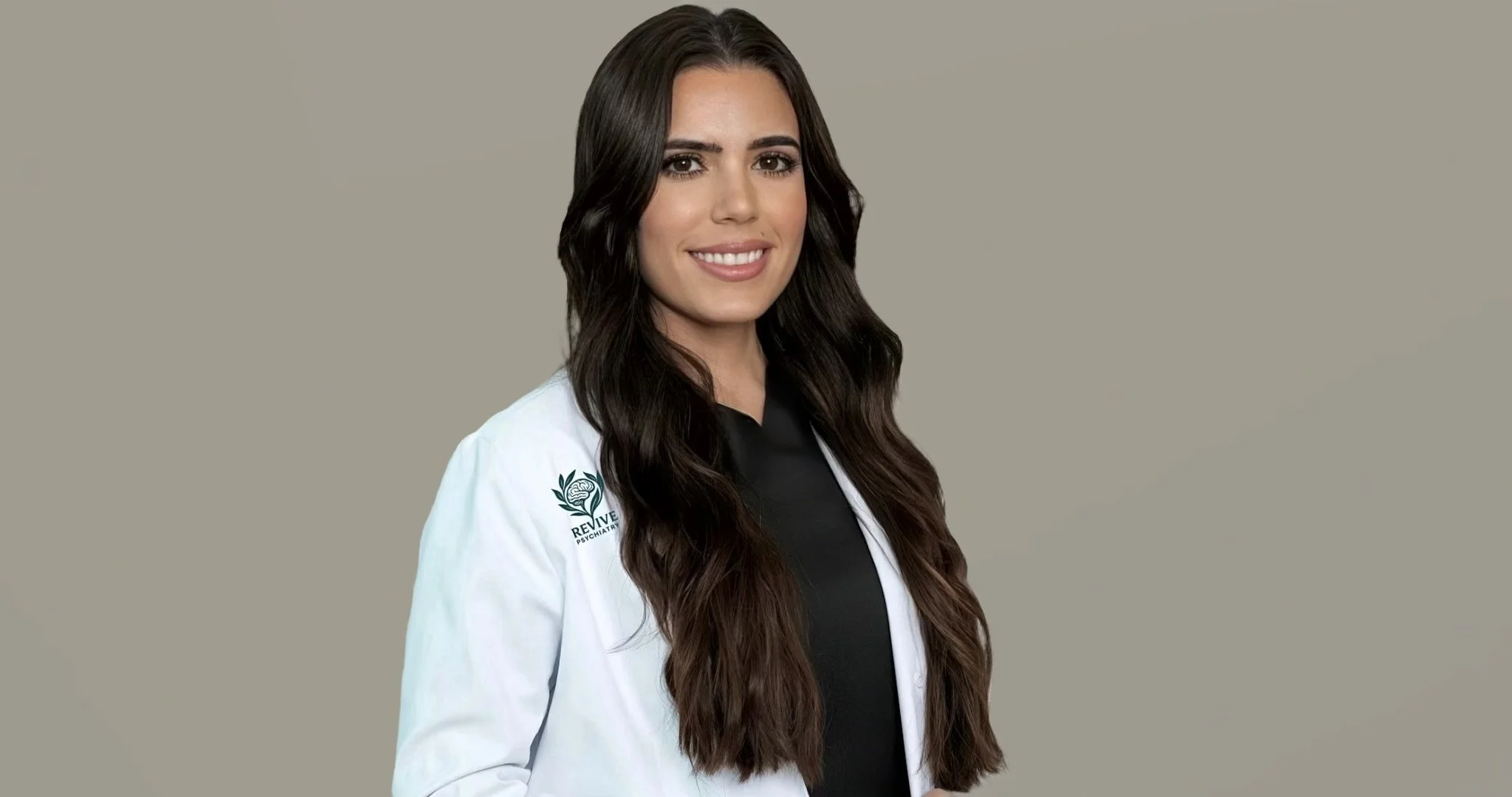 A woman with long dark wavy hair wearing a white lab coat with a logo on the sleeve, smiling against a neutral background.