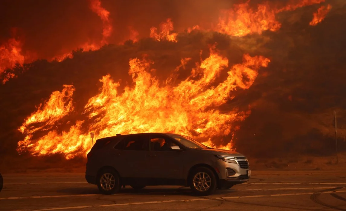 A car on the side of a road with a wildfire burning on the hillside behind it.