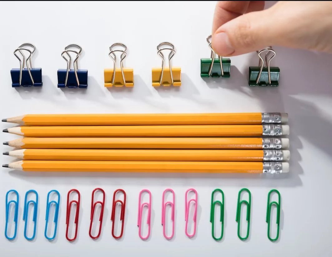 Eight yellow pencils with erasers, six paperclips in blue, red, pink, and green, and six binder clips in yellow, green, and blue on a white surface. A hand holds a green binder clip.