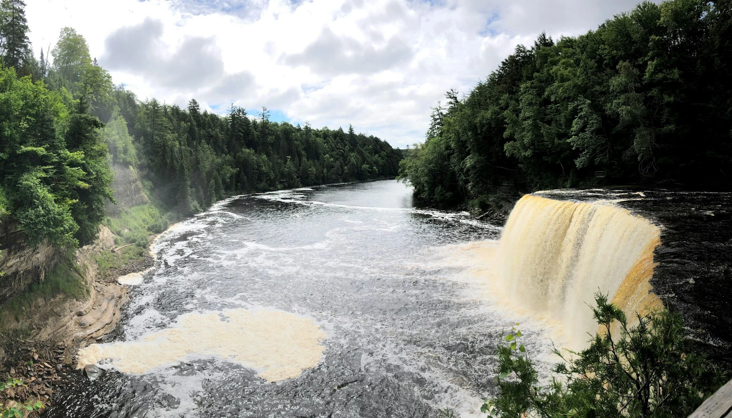 A river with a waterfall surrounded by green trees under a cloudy sky.