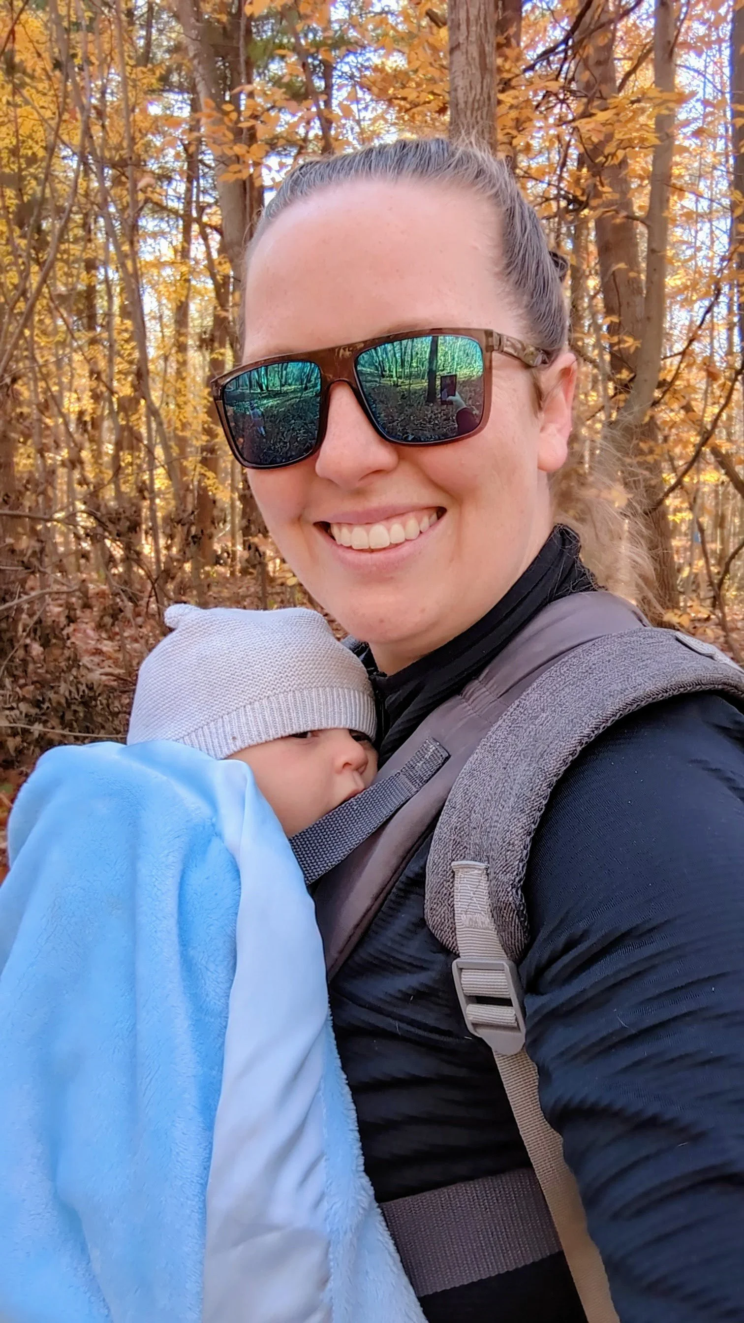 A woman with sunglasses smiling and carrying a baby with a grey hat in a baby carrier, walking in a wooded area with orange and yellow autumn leaves.