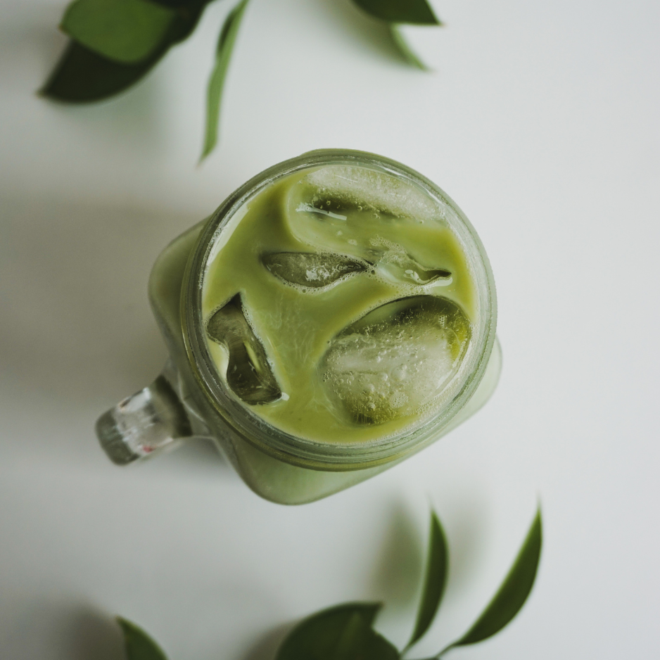 A glass jar filled with green matcha latte and ice cubes, viewed from above, on a white surface with green leaves around it.