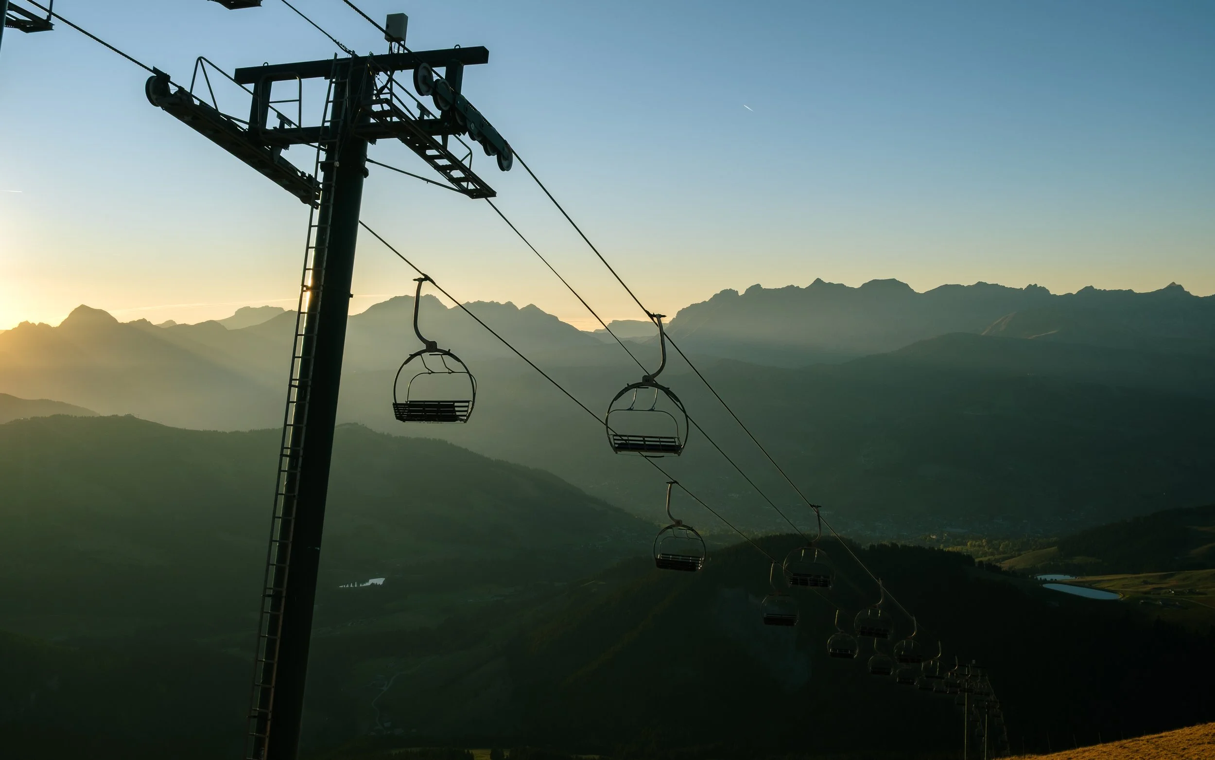 Empty ski lift chairs suspended on a cable in a mountainous landscape at sunset or sunrise, with a clear sky.