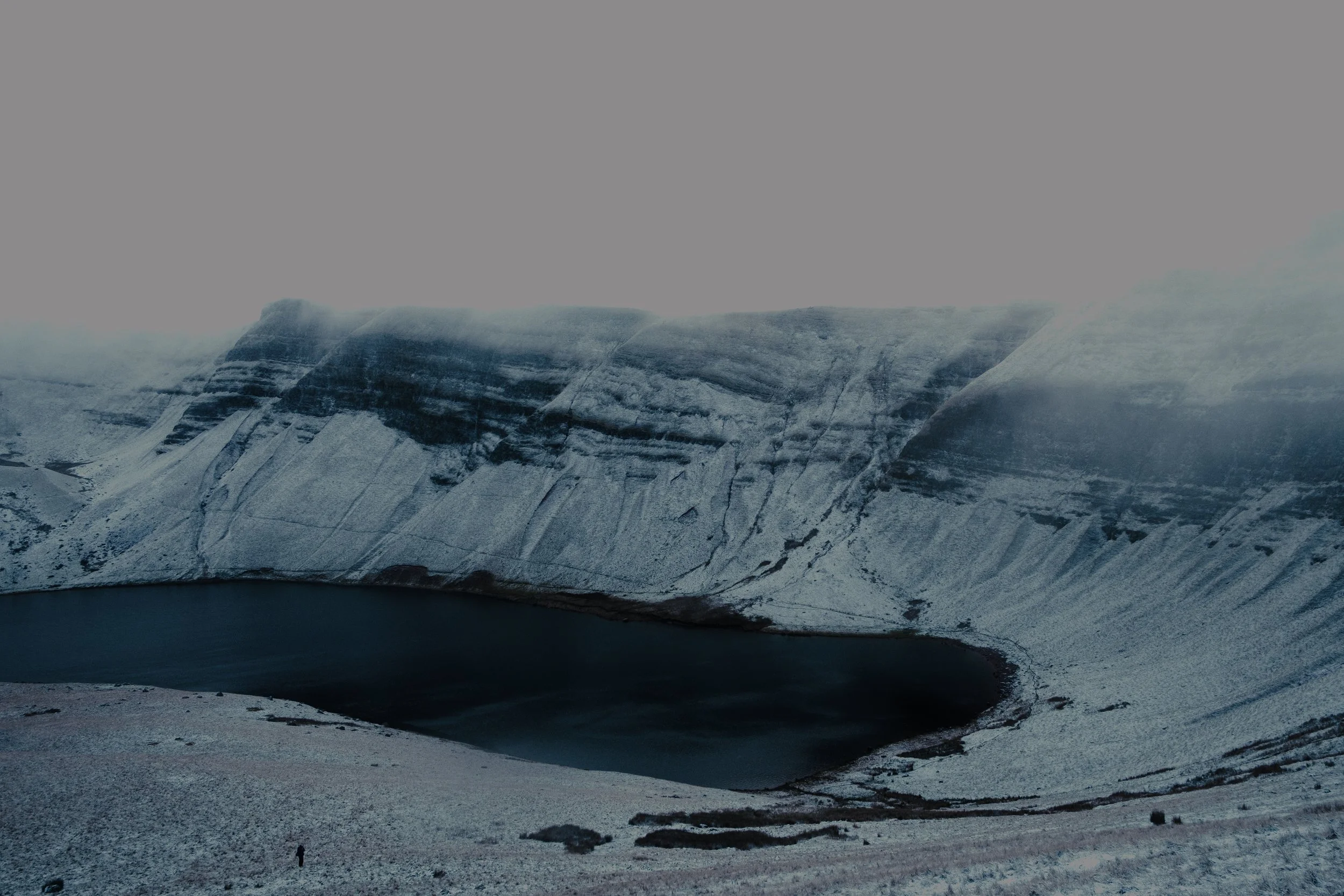 Snow-covered mountain with a crater lake in the foreground, partially shrouded in fog.