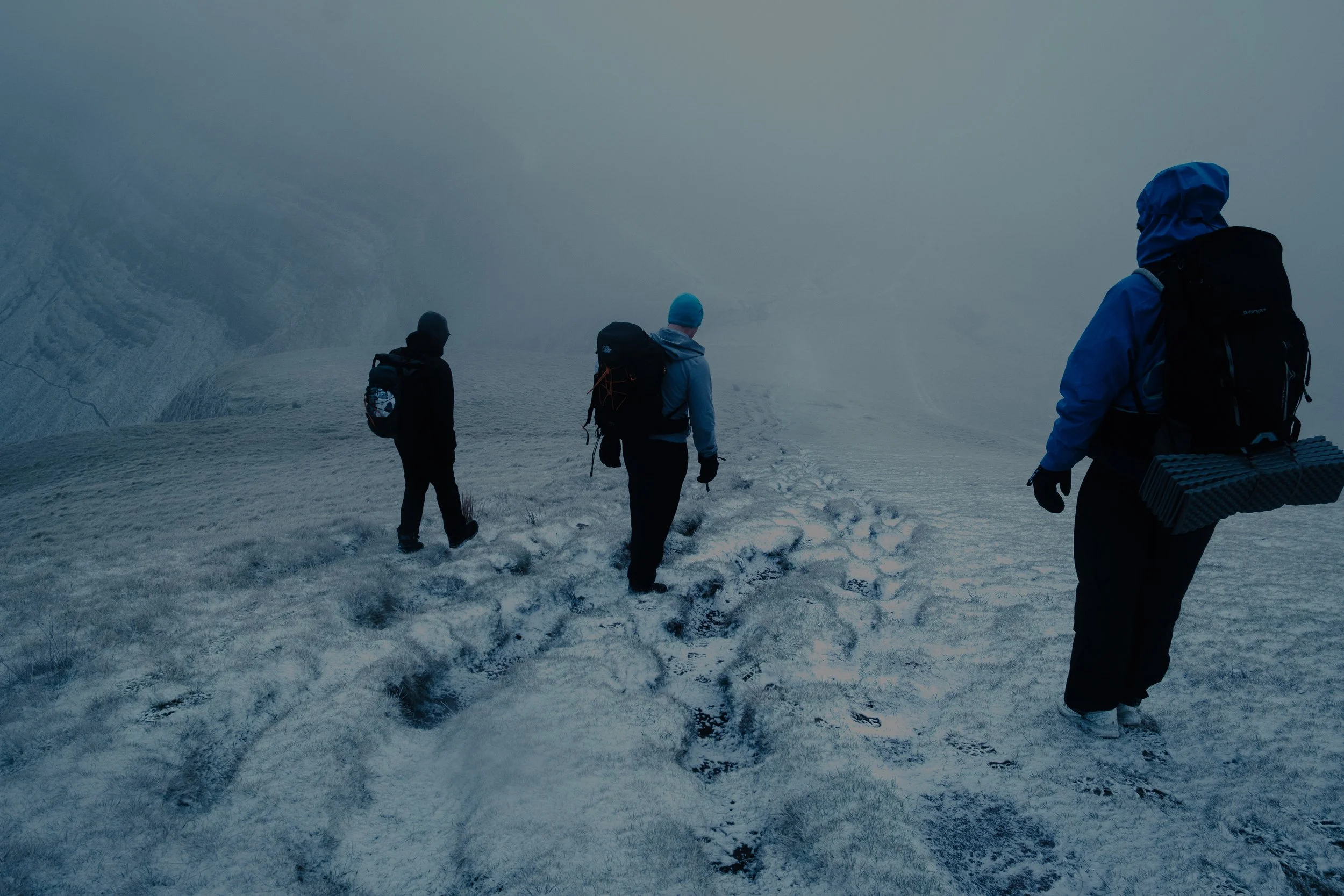 Four hikers walking in snowy, foggy mountain terrain with backpacks and outdoor gear.