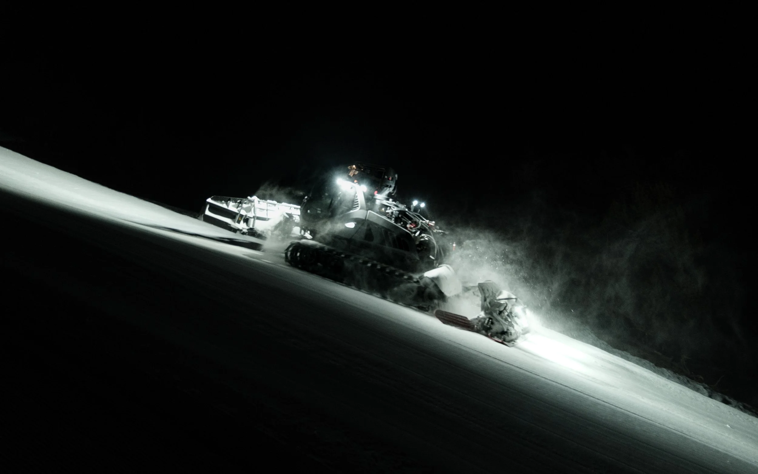 Nighttime image of a snowcat vehicle plowing snow on a slope, with snow flying around it.