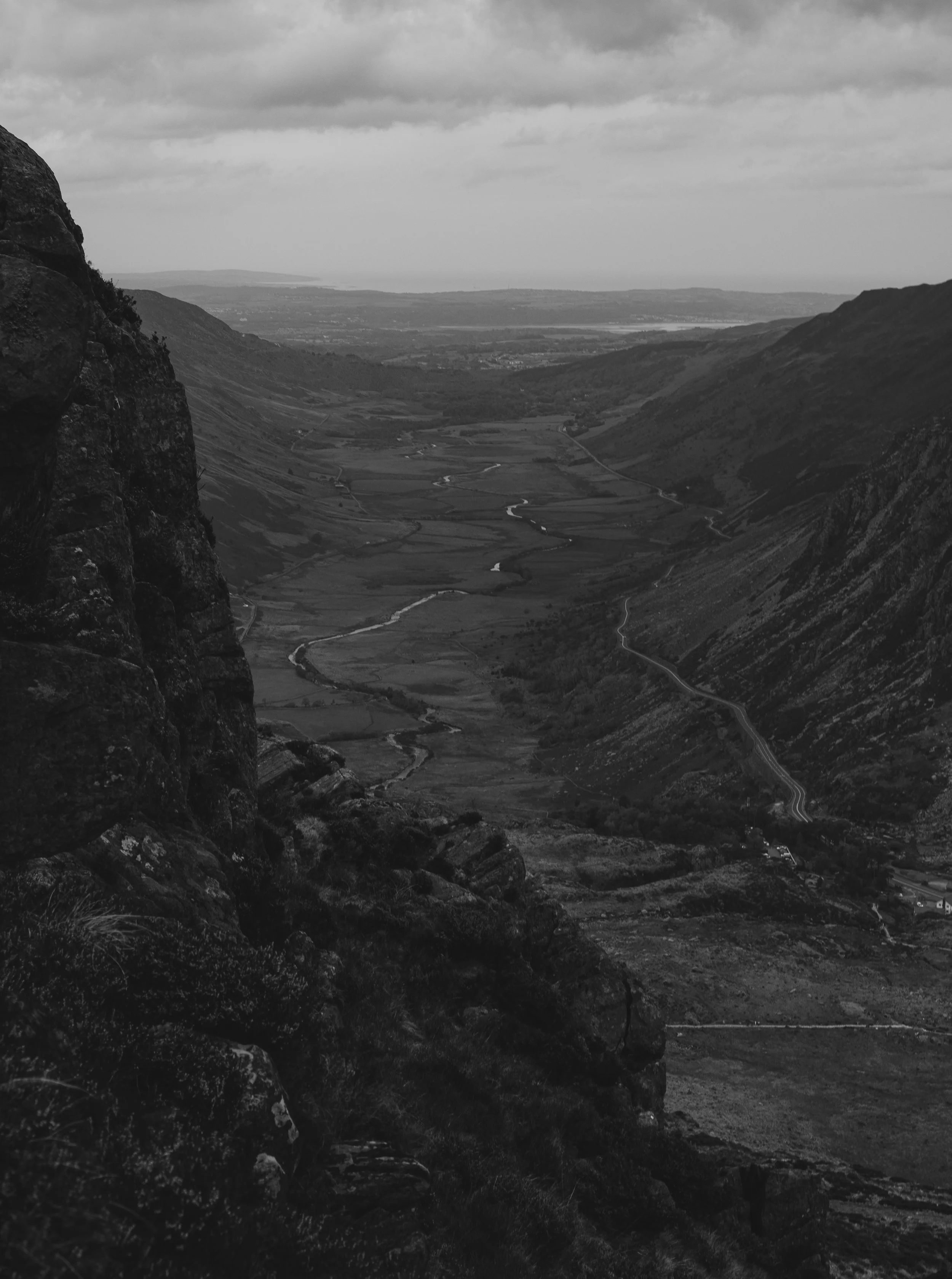 A black and white landscape photograph of a valley surrounded by mountains, with winding streams and a distant body of water under a cloudy sky.