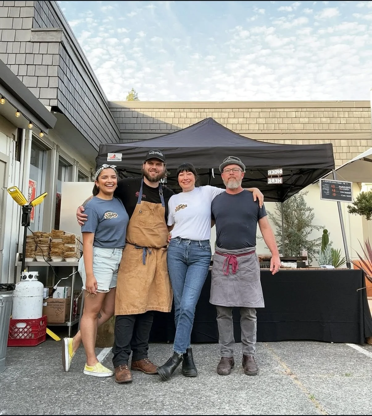 Four people standing in front of a food stand, smiling and posing for a photo. Two women and two men, some wearing aprons, likely working at the food stand, with outdoor setting and a building in the background.