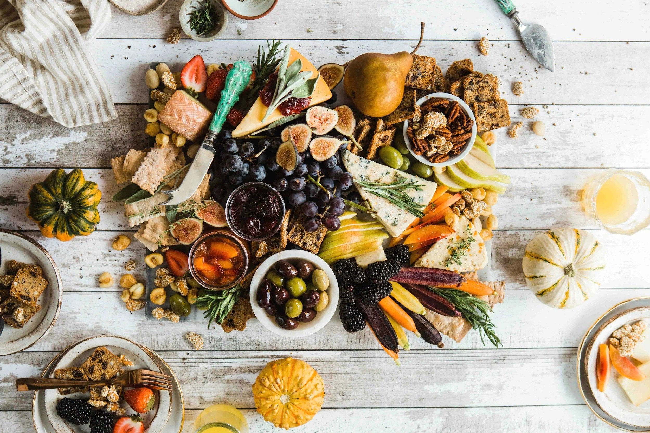 A beautifully arranged cheese and fruit platter with grapes, blackberries, strawberries, figs, and a variety of cheeses on a white wooden table, accompanied by small pumpkins and glasses of yellow beverage.