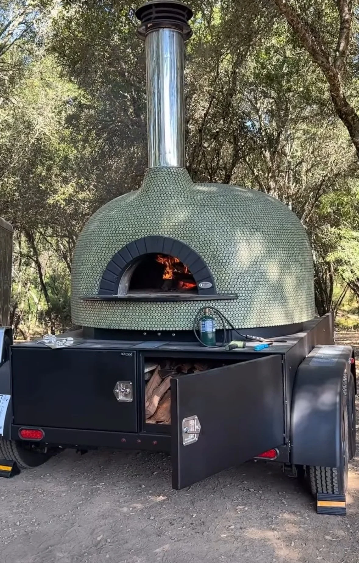 A mobile wood-fired pizza oven on a trailer, with a dome-shaped oven exterior, a chimney on top, and fire visible inside the oven, set outdoors among trees.
