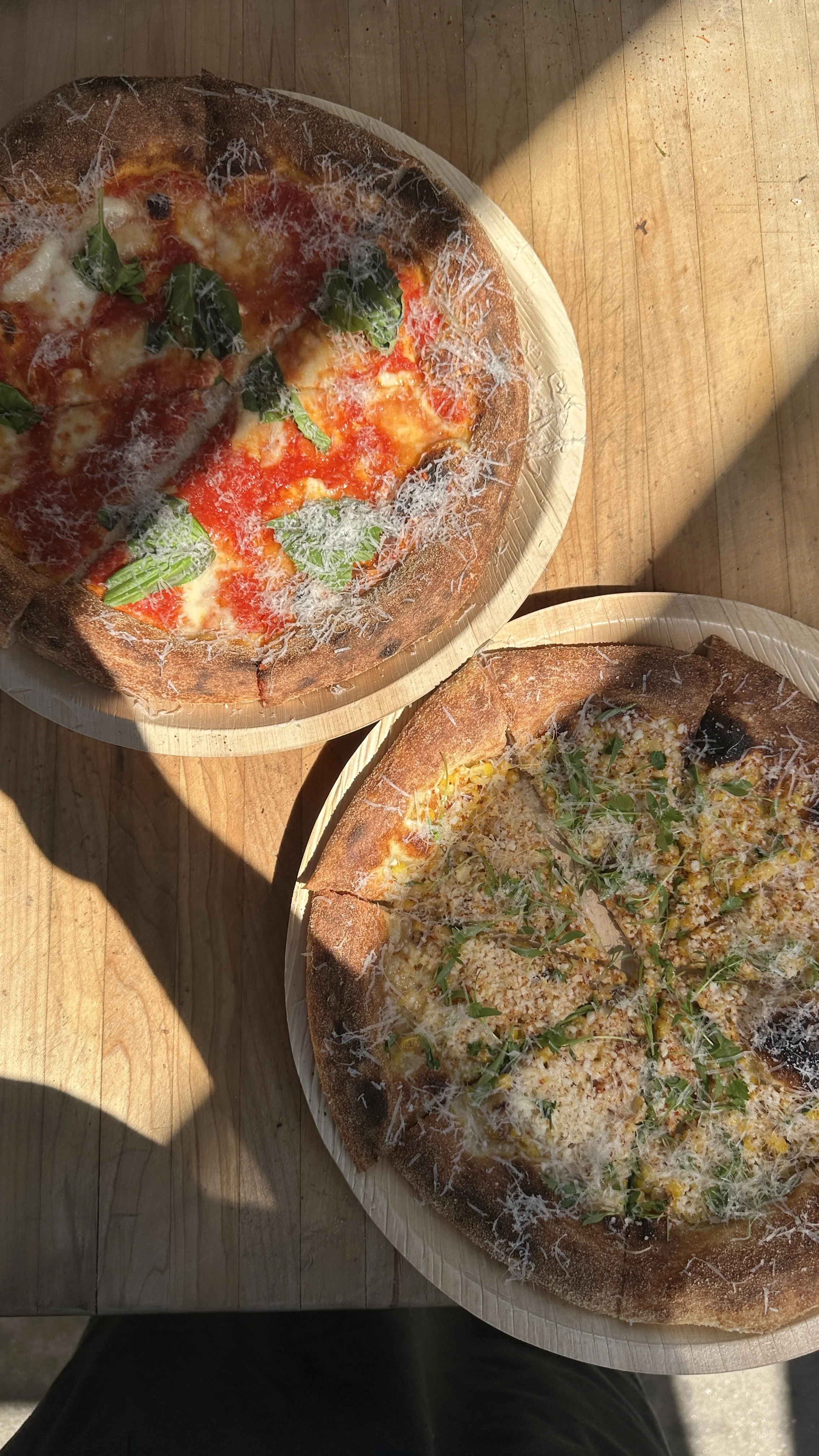 Two pizzas on wooden trays on a wooden table, with a shadow cast across part of the table and pizzas. One pizza has tomato, cheese, basil, and grated cheese toppings. The other pizza has cheese, arugula, and grated cheese toppings.