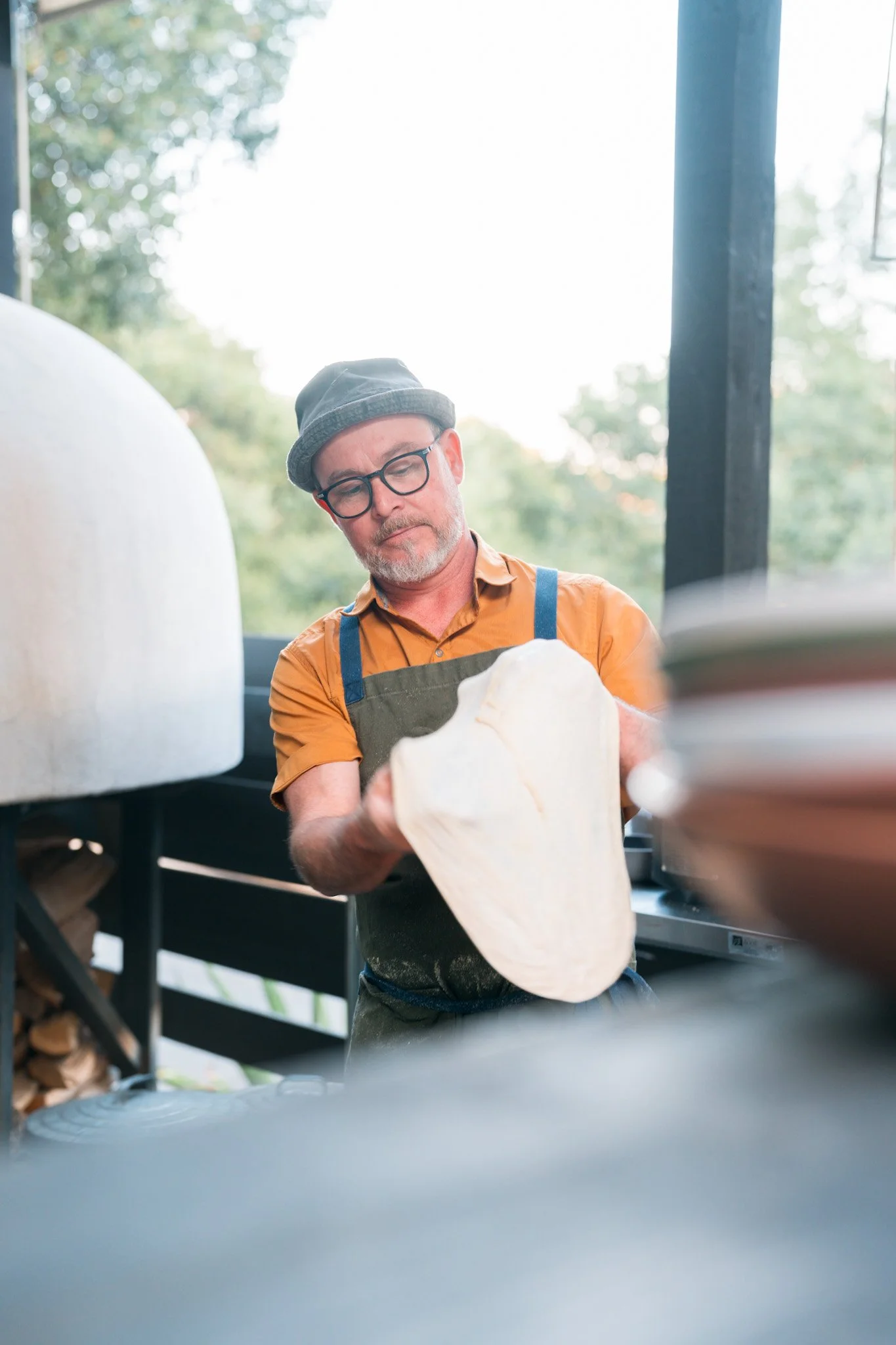 A man wearing a hat, glasses, and an apron preparing dough in a kitchen or outdoor area with trees visible through the window.