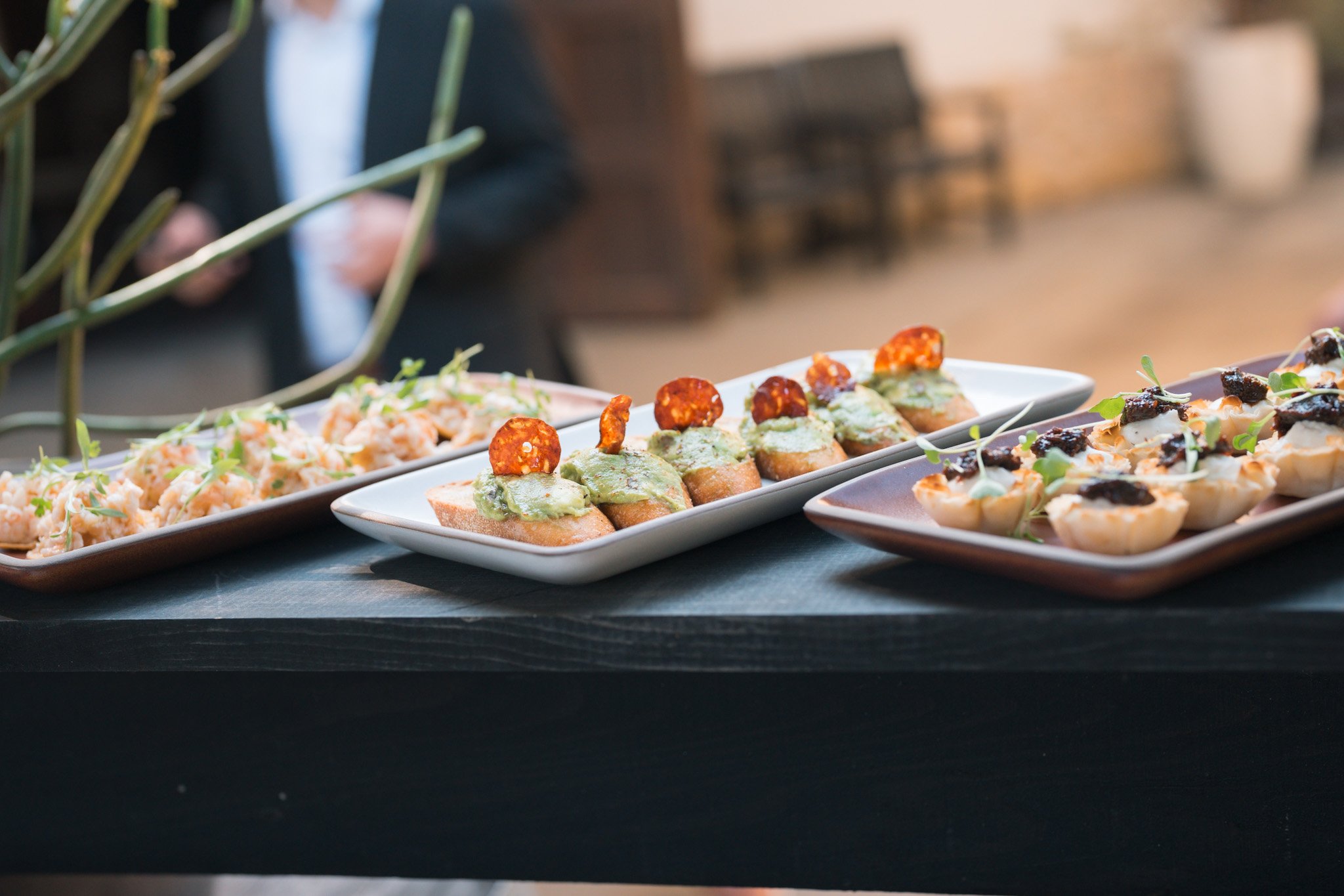 Appetizers on white and brown rectangular plates with shrimp, toast with guacamole and sliced pepperoni, and deviled eggs garnished with microgreens on a black table.
