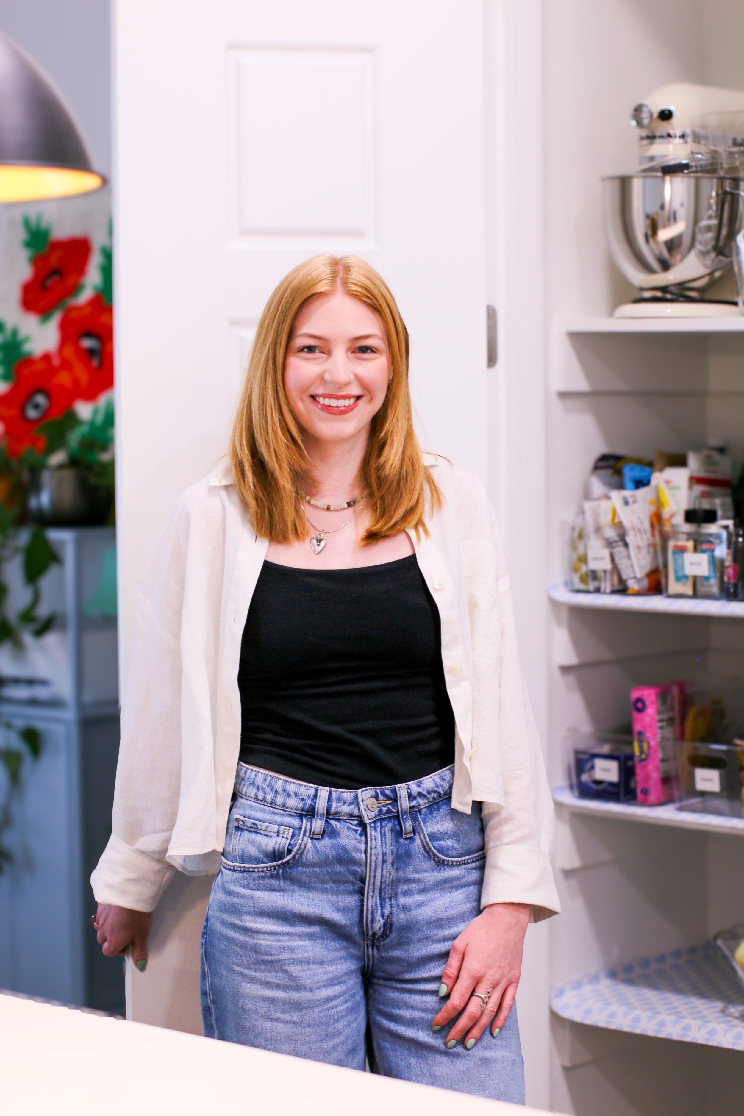 A woman with long reddish hair smiling, wearing a black top, light-colored cardigan, and jeans, inside a store with shelves of various products in the background.