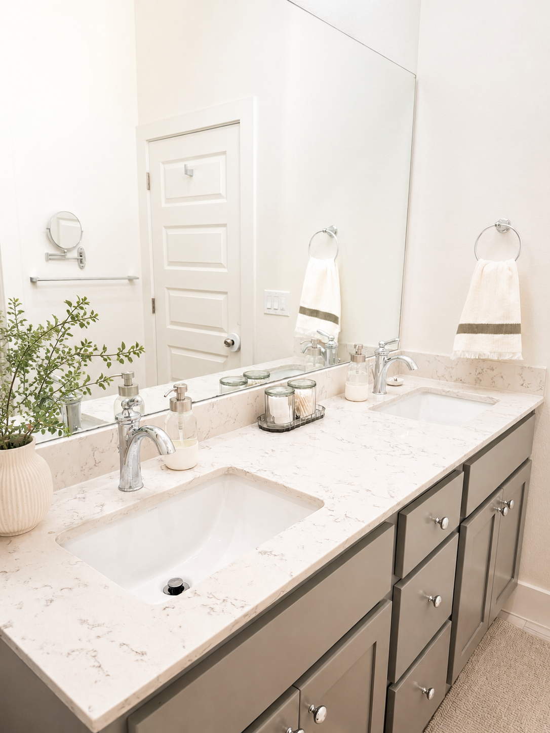 Modern bathroom with dual sinks, marble countertop, large mirror, towels on ring holders, small potted plant, and bathroom accessories.
