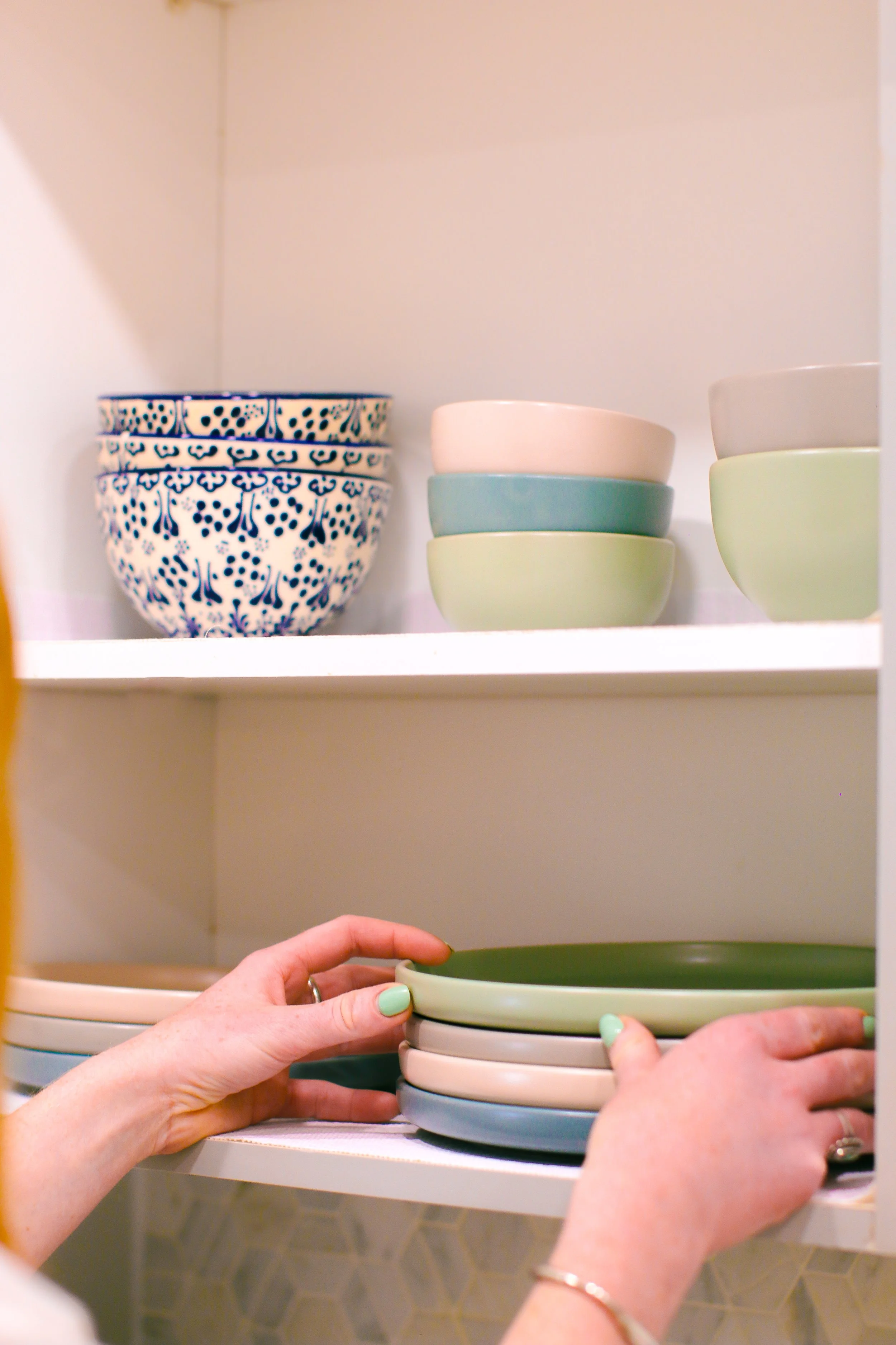 Person organizing a stack of pastel-colored bowls on a kitchen shelf. The bowls are in shades of green, blue, white, and beige, with decorative patterned bowls on the top shelf.